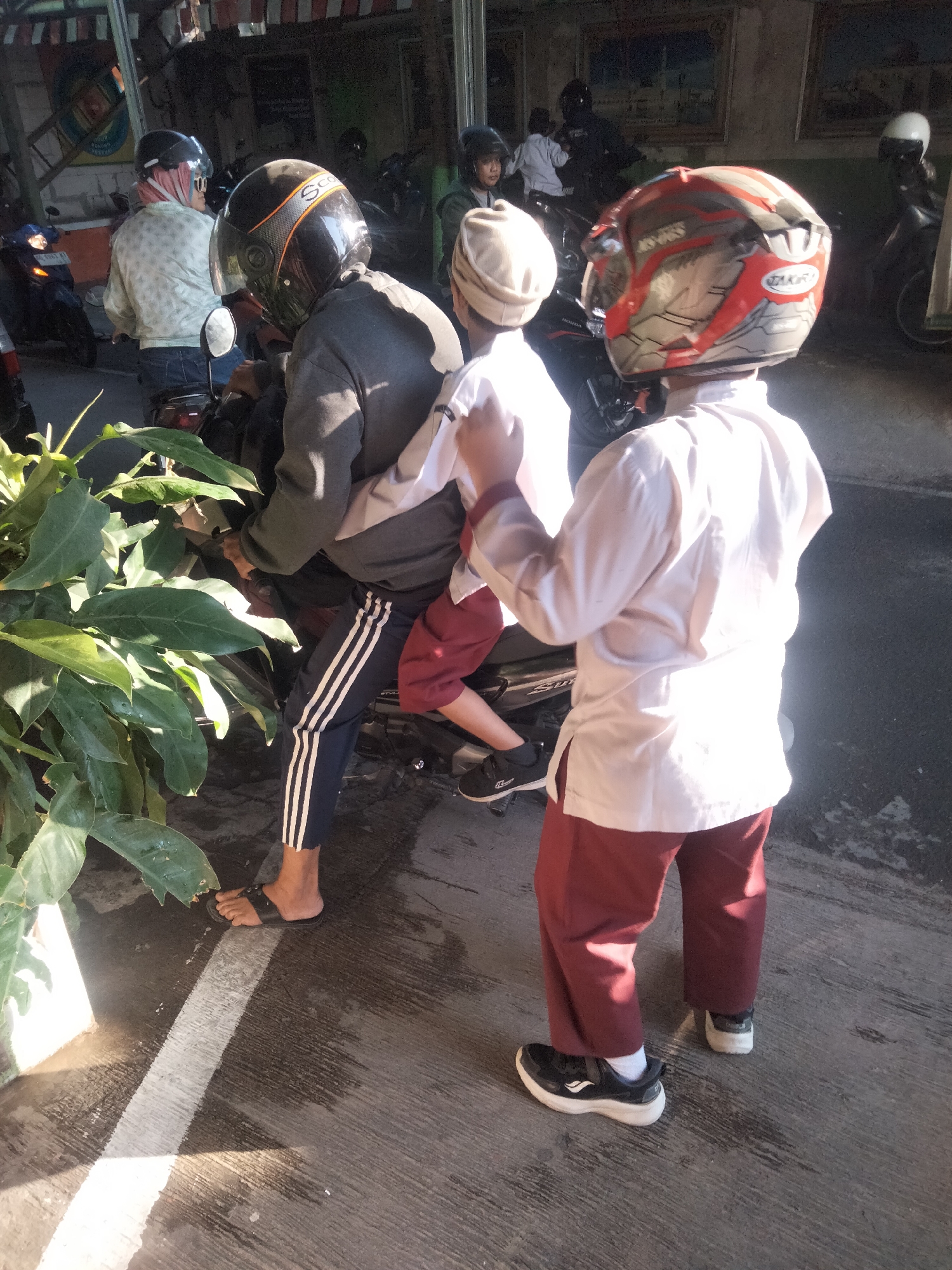 The image depicts a street scene in Surakarta, Indonesia, likely near a commercial area given the awnings and storefronts visible in the background. The foreground is dominated by two young boys on a motorcycle. The boy in the front wears a red and silver helmet, a light-colored shirt, and dark red pants. The boy behind him has on a light-colored cap, a dark gray jacket, and dark pants with white stripes. Both are wearing helmets, indicating adherence to local traffic laws.
The motorcycle is parked on a paved road, with a large, leafy plant to the left of the frame. Sunlight casts strong shadows, suggesting it is daytime. In the background, other motorcycles and people are visible, some appearing to be vendors or shoppers. The architecture in the background includes what appear to be shops with striped awnings and framed artwork on the walls, typical of urban Indonesian settings. The overall impression is a bustling, everyday street in Surakarta.