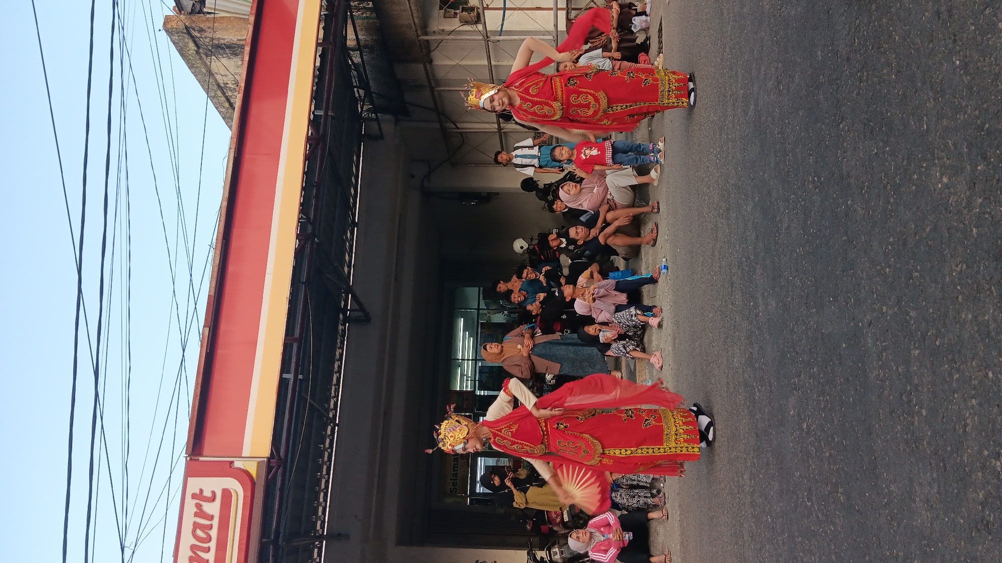 The image depicts an outdoor street performance in front of a building with a red and yellow awning that says "mart". Two individuals in traditional red and gold costumes, adorned with ornate headpieces and elaborate makeup, are performing a dance. One performer on the left is holding a red fan. A crowd of spectators, dressed in casual clothing, are seated on the ground watching the performance. The setting appears to be a street in Tanjung Anom, Indonesia, as evidenced by the architecture and the presence of motorbikes. The sky is clear blue with power lines overhead. The ground is paved with asphalt.