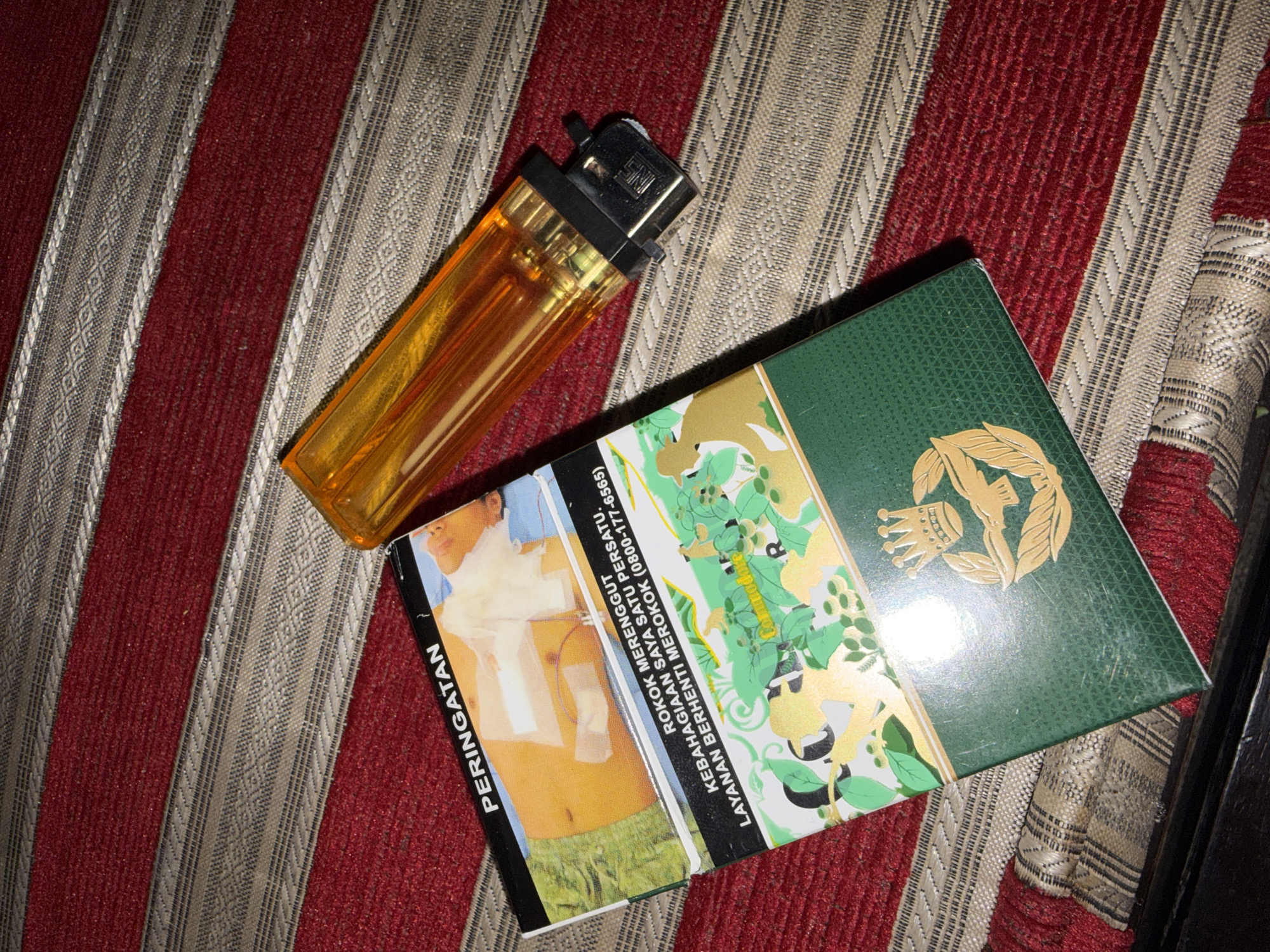 An overhead shot shows a pack of cigarettes and a bright orange lighter on a red and beige striped surface. The cigarette pack is partially open, revealing a graphic image on the front warning about the dangers of smoking. Text on the pack, written in Malay, reads "PERINGATAN" (Warning) and "ROKOK MERENGGUT KEBAHAGIAAN SAYA SATU PERSATU. LAYANAN BERHENTI MEROKOK (0800-177-6565)" which translates to "Smoking steals my happiness one by one. Quit Smoking Service (0800-177-6565)". The bottom part of the pack displays a green design with gold embellishments, including a crown and laurel wreath. The lighter is positioned to the right of the cigarette pack, with its transparent orange casing revealing the internal mechanism. The striped surface appears to be fabric, possibly a rug or upholstery. The lighting suggests an indoor environment, with the angle of the light creating some shadows. The specific location in the city of Tupah, Indonesia, is not discernible from the visual information provided.