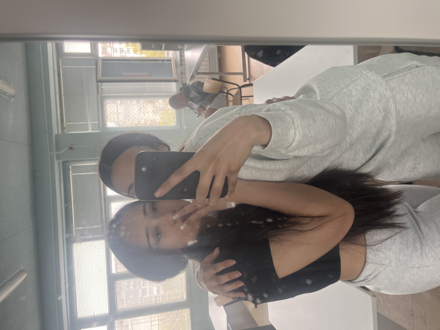 A mirror selfie captures two young women in what appears to be an indoor setting in Sofia, Bulgaria.
The primary subject is a woman with long, dark hair, positioned centrally in the mirror's reflection. She is wearing a black top and light grey sweatpants, and her arms are around another person. Her face is visible, looking towards the camera, with a neutral to slight smile. Her fingernails are visible and appear to have light-colored polish.
Embracing her from behind is a second woman, who is holding a dark-colored smartphone to take the mirror selfie. Only her upper face, including her eyes and forehead, is clearly visible above the phone. She is wearing a light grey hooded sweatshirt. Her hand holding the phone shows a ring on one finger.
The background, seen through the reflection, reveals a room with large windows that have horizontal blinds. Bright daylight is entering through the windows, suggesting it is daytime. Outside the windows, some greenery and buildings can be vaguely seen. Inside the room, beyond the women, there are light-colored walls and what appear to be tables or desks. Another person, partially visible and blurred, is seated further in the background near a table, suggesting a common area, classroom, or cafeteria. No specific text is visible within the image. The overall atmosphere appears casual and friendly.