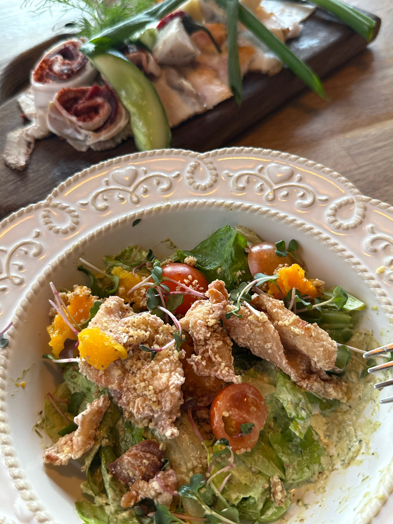 An overhead, close-up shot captures a white, scalloped-edge bowl filled with a vibrant salad. The salad is a mix of fresh green lettuce, sliced cherry tomatoes, bright orange segments of mandarin oranges, and pieces of crispy, breaded chicken. Microgreens add a delicate touch throughout. The bowl sits on a wooden table, with another wooden board blurred in the background, holding a selection of meats and a slice of cucumber, garnished with dill. The lighting suggests an indoor setting, likely a restaurant.