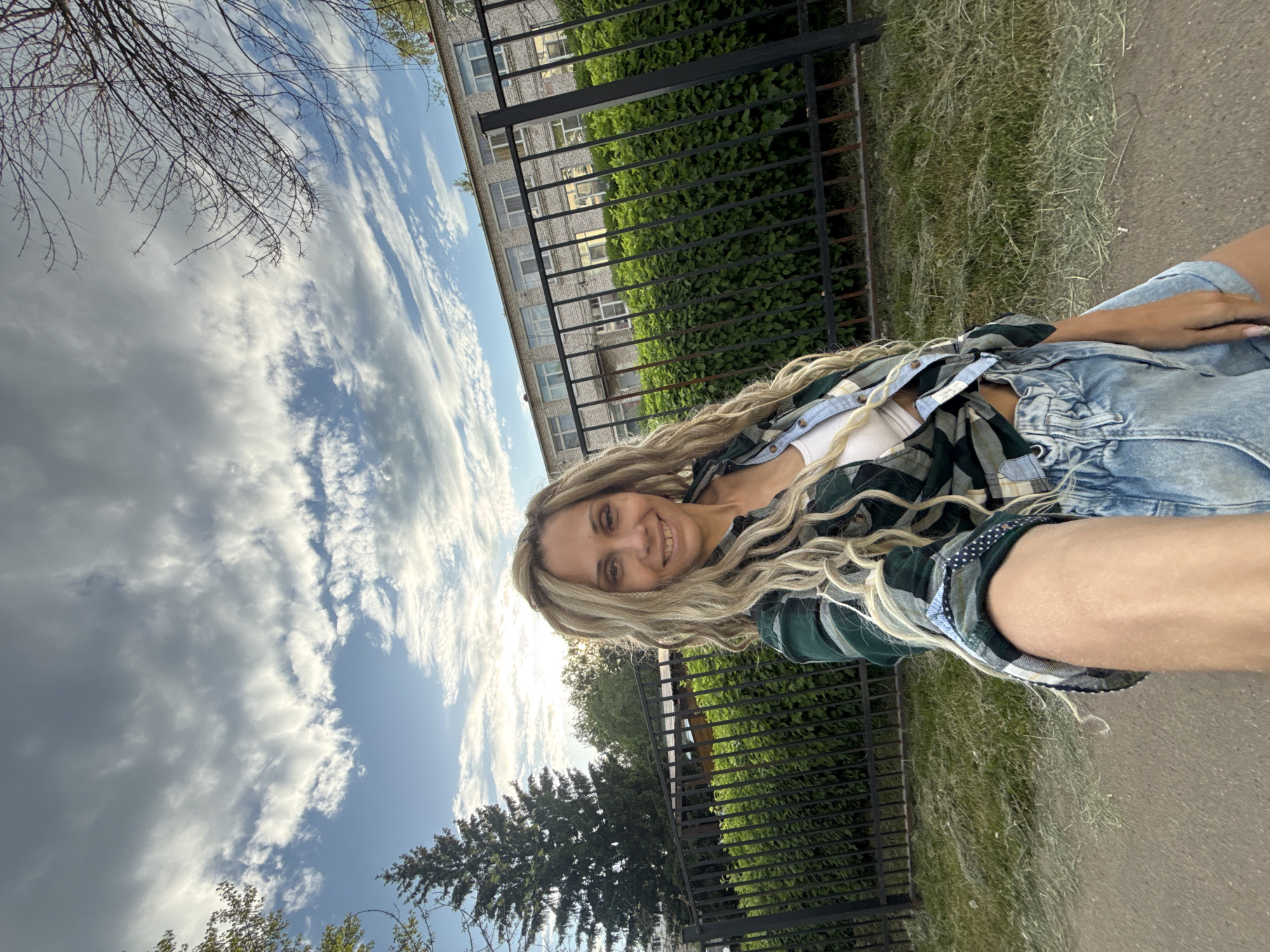 A young woman poses for a photo outdoors. She has long, wavy blonde hair and is wearing a white top, a green and black plaid shirt, and a denim jacket. She is smiling at the camera.
Behind her, there is a black metal fence with green foliage growing behind it. Further back, a multi-story building with light-colored walls and dark window frames is visible. To the left of the building, there are bare tree branches against a cloudy sky.
On the other side of the fence, there is a large evergreen tree. The ground is paved.
While the location is stated to be Rusanovo, Russia, there is no specific detail within the image to definitively confirm this.