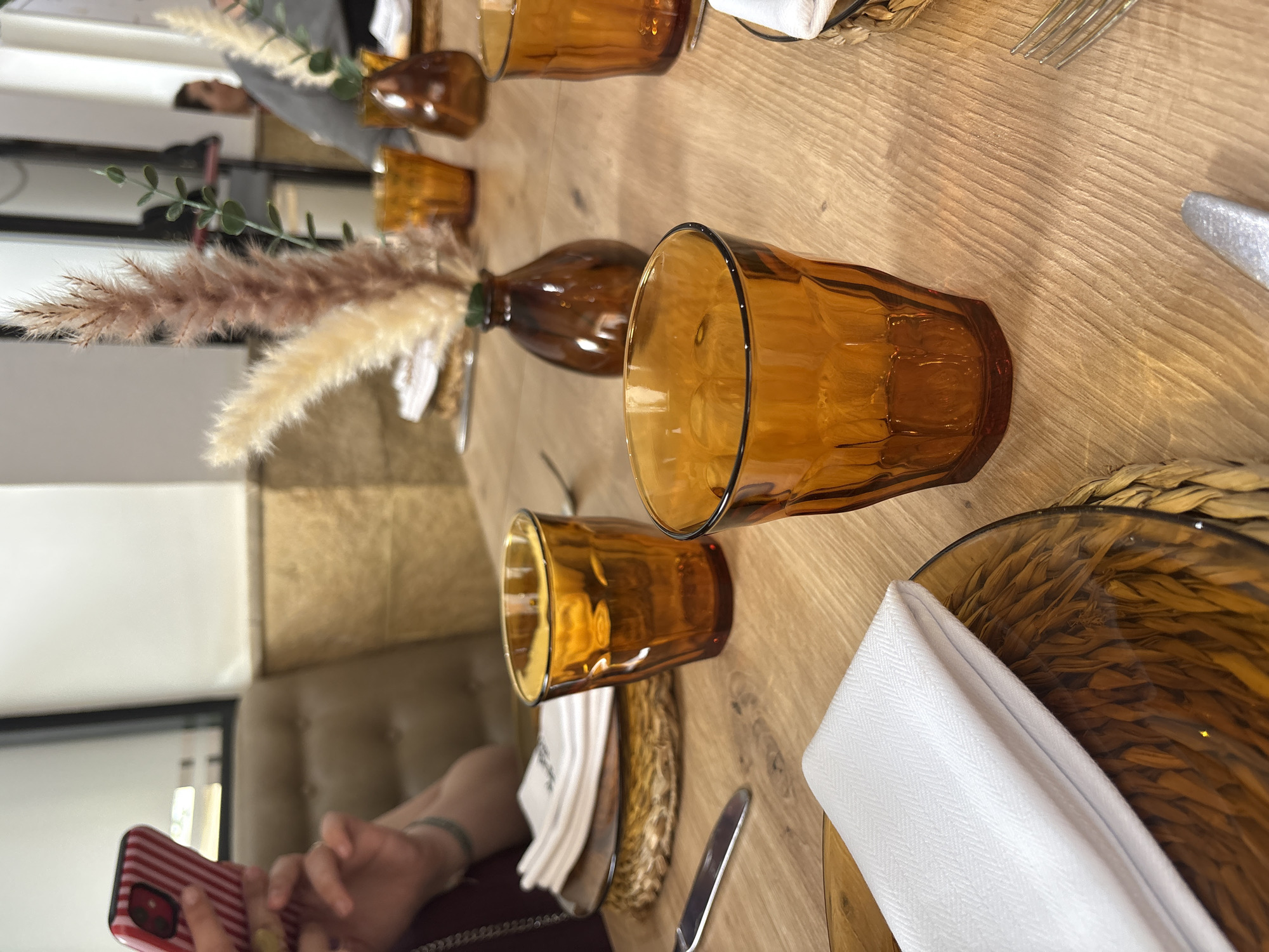 A table setting with amber-colored glasses, wicker placemats, white napkins, and cutlery. A floral arrangement in amber glass vases adorns the table's center. A person holds a red-and-white striped phone. The location is most likely in the city of Córdoba, Spain.