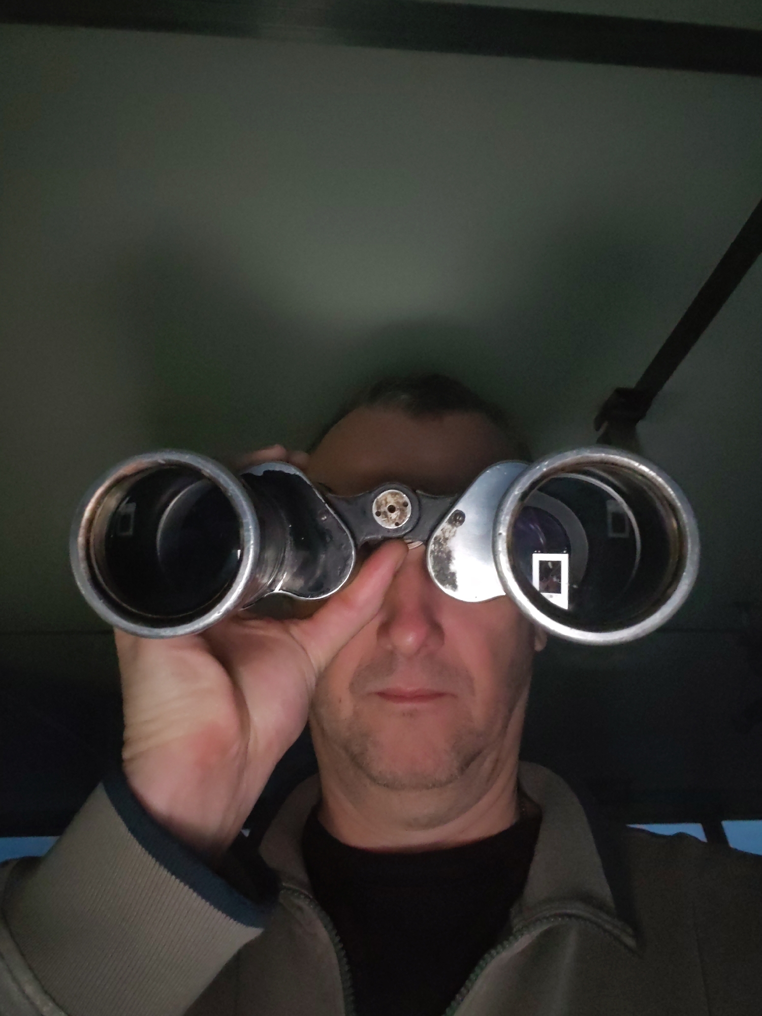 A middle-aged Caucasian man is viewing through binoculars, held up to his eyes with his left hand. The binoculars are vintage, silver-colored, and show reflections of light within the lenses. The man is wearing a dark-colored shirt beneath a zipped-up khaki-colored jacket with a dark blue trim on the sleeve. He has short, receding gray hair. The background appears to be the interior of a vehicle with a greenish-gray ceiling and a dark-colored bar or handle extending from the ceiling. The location is not discernible as Bereg, Russia based solely on visual information in the photo.
