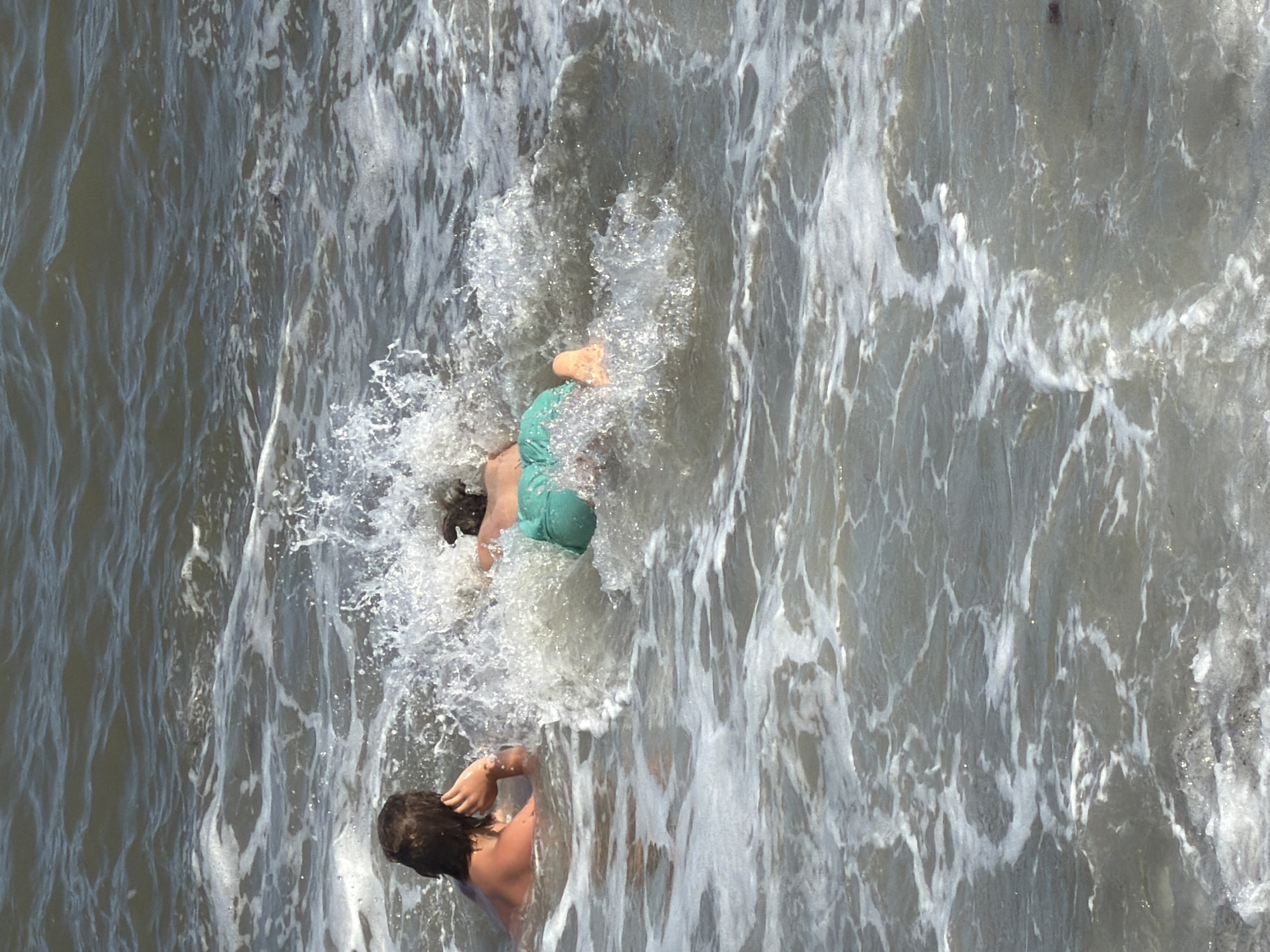 Two individuals are swimming in the sea at Alushta, Ukraine. One person, seen from the back, is lying on their stomach with their head resting on their hand. The other person is in front, in a diving position, with their legs in the air and kicking as they submerge themselves in the water. The scene is dynamic, with waves crashing and creating splashes around the swimmers. The water is a mixture of dark blue and white foam, indicating the movement of the waves.