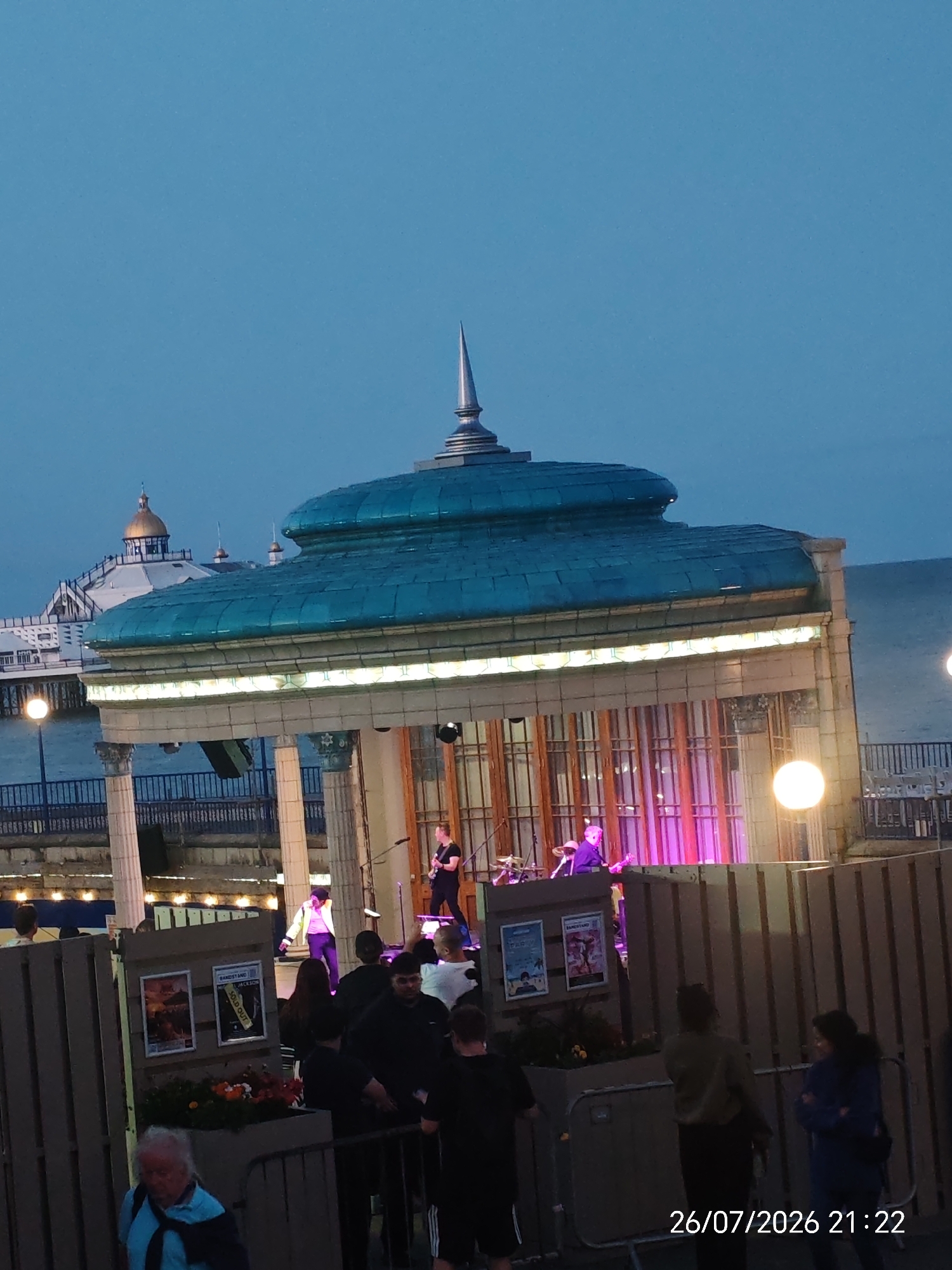 This image captures a twilight scene at the Eastbourne Bandstand, East Sussex, United Kingdom. The focal point is the bandstand's distinctive turquoise roof, topped with a silver spire. A band is performing on the illuminated stage, with musicians playing guitars and drums. The stage area is lit with vibrant pink and purple lights, creating a festive atmosphere. In the background, the iconic Eastbourne Pier can be seen extending into the sea, with its white structure and golden dome.
Several people are visible in the foreground, some standing behind barriers, seemingly watching the performance. One person in a bright yellow jacket and dark trousers stands out on stage. The environment suggests an outdoor event on a pier or promenade, with the sea visible to the right. The overall impression is of a lively evening entertainment event. The timestamp "26/07/2026 21:22" is overlaid on the bottom right corner.