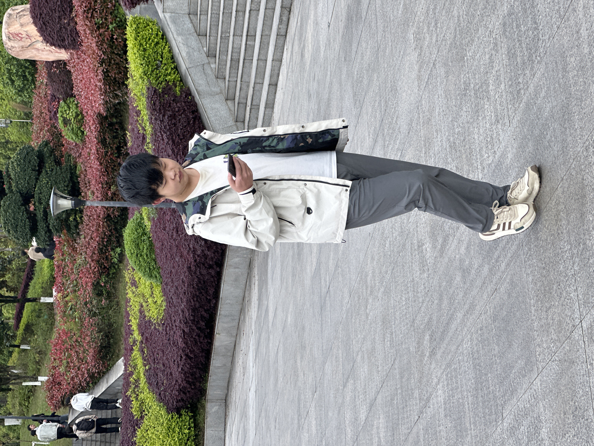 A person stands in an outdoor area in Jiangjin, China. The individual wears gray pants, a white t-shirt, and a light-colored jacket with a patterned interior. The person is looking at a mobile phone held in their hands. They also have on sneakers. They are standing on a gray-tiled surface. In the background, there are stairs, various types of bushes with colorful foliage, and a light post. Other people are visible further in the distance.