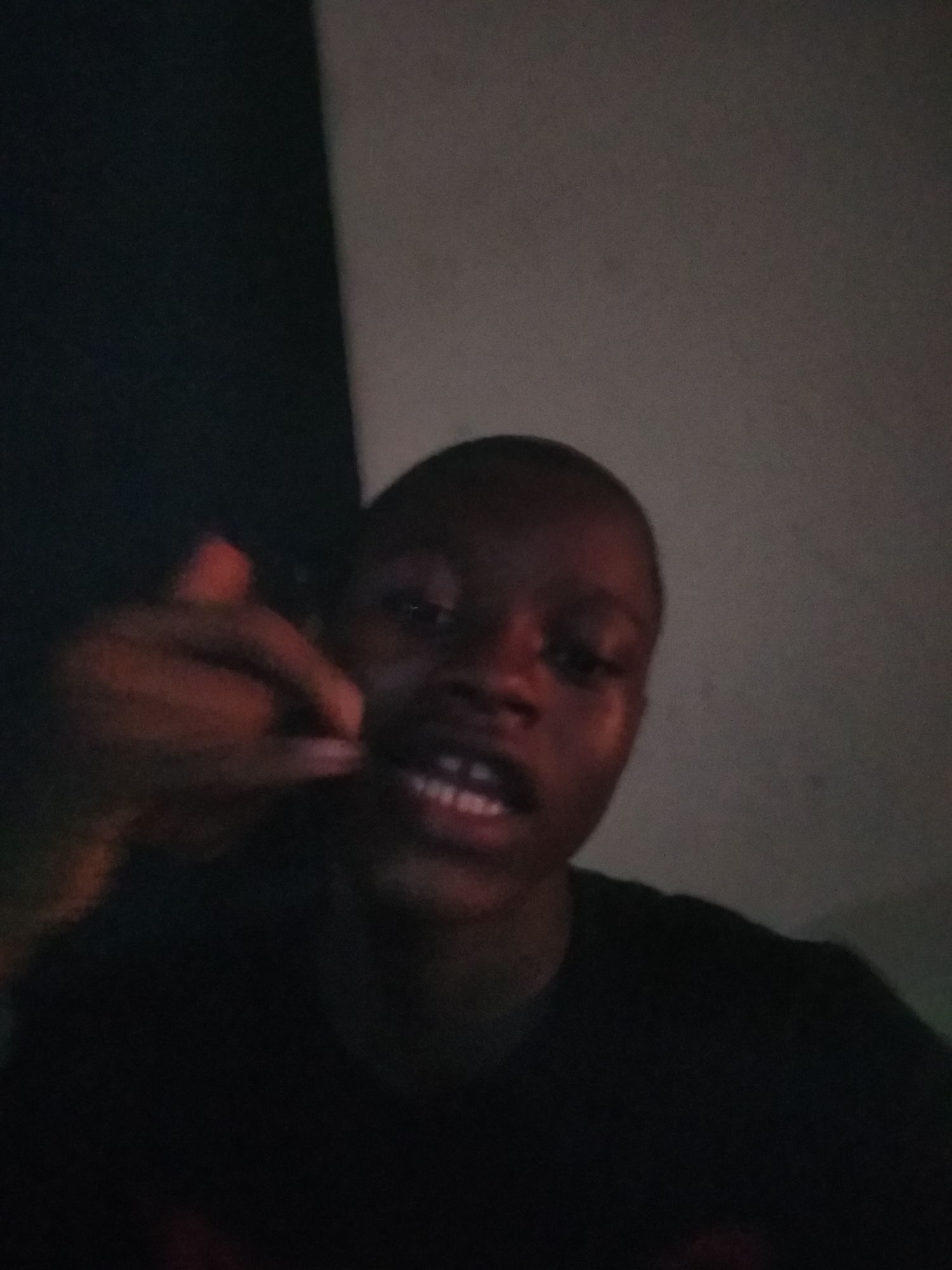 This low-light, close-up portrait features a young person, likely in an indoor setting in Shangisha, Nigeria. The individual, with dark skin and short hair, is positioned centrally, looking directly towards the camera. Their face is partially illuminated, highlighting their open mouth which reveals visible white teeth, suggesting they might be speaking, smiling, or singing.
To the left of their face, their right hand is raised, making a gesture where the thumb and pinky finger are extended while the middle fingers are curled inwards, commonly known as a "call me" sign. The hand appears slightly blurred, possibly due to motion or the challenging lighting conditions.
The background is simple and indistinct, divided into two main areas: a dark, almost black section on the left, and a lighter, muted grey-brown section on the right, indicating a corner or a junction of surfaces. The overall environment is poorly lit, giving the image a grainy texture typical of low-light photography. There is no visible text. The informal nature of the pose and the dark setting suggest a casual moment, possibly at night or in a dimly lit room.