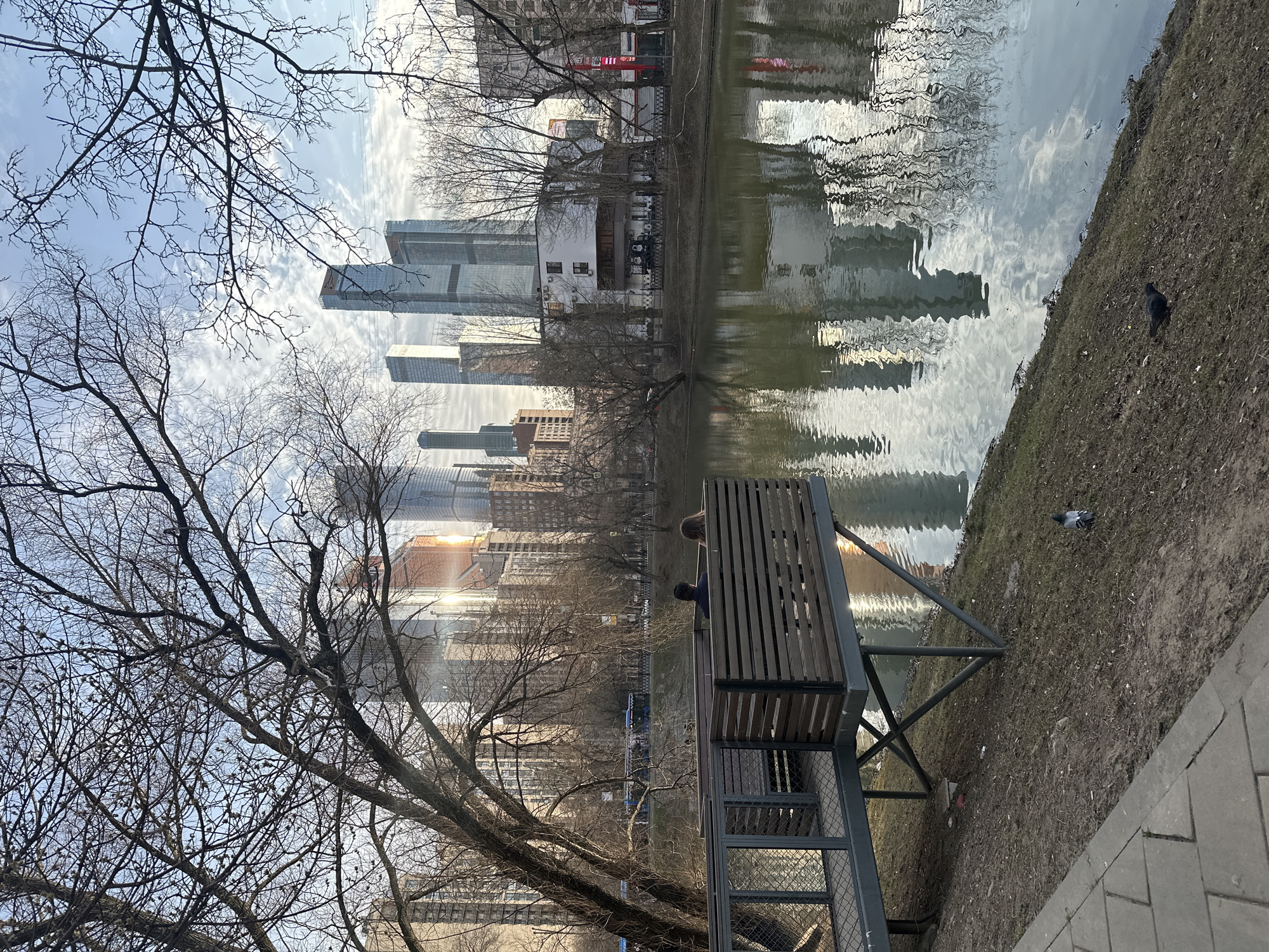 The image depicts an outdoor urban landscape in Moscow, Russia, specifically featuring Presnensky Pond with the prominent skyscrapers of the Moscow International Business Center (Moscow City) visible in the background. The scene is illuminated by late afternoon sunlight, causing glints on the buildings and reflections on the water.
In the immediate foreground, a paved pathway runs along a sloped bank of bare earth and sparse vegetation adjacent to the pond. Two pigeons are observed on this bank: one dark-colored pigeon near the pathway on the left, and another darker avian subject further to the right by the water's edge.
An elevated structure, consisting of a metal-grated walkway leading to a wooden platform with horizontal slatted barriers, extends over the pond from the left. This platform is supported by dark metal beams. Two individuals are seated on the platform, facing away from the camera, looking towards the pond and the distant city skyline. The person on the left is dressed in dark attire, while the person on the right has lighter-colored hair and wears a lighter-toned jacket.
The pond's surface is calm, reflecting the partially cloudy blue sky, scattered clouds, and the vertical structures of the distant city. On the far bank of the pond, bare deciduous trees line a road. A blue public transport bus is partially visible on this road. Beyond the road, several low-rise buildings are present, including a white two-story building with dark windows and a red sign on the right. The background is dominated by numerous modern glass and steel skyscrapers characteristic of Moscow City. Bare tree branches partially frame the upper and left portions of the image.