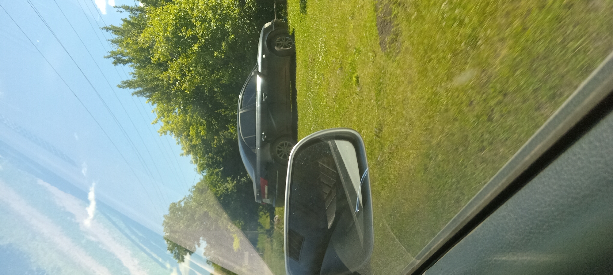 A black sedan is parked on a grassy area next to a line of trees under a clear blue sky with a few scattered clouds. The car is seen from the side, with its rear facing towards the right of the frame. A side-view mirror is visible in the foreground on the left, reflecting a portion of the car's interior. The scene appears to be on the outskirts of Koviagy, Ukraine.