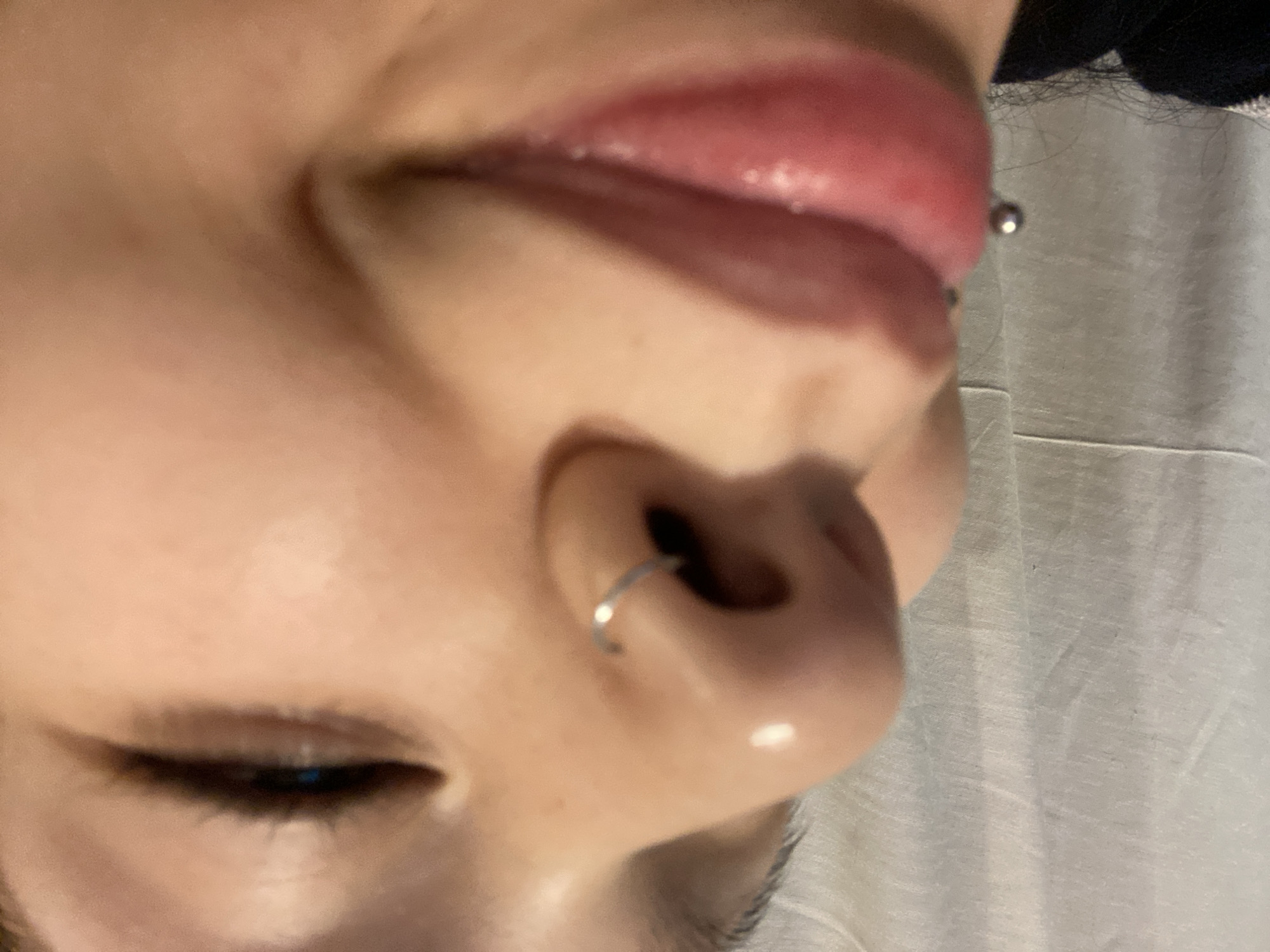Close-up of a person's face, focusing on the nose, mouth, and eye. The person has a septum piercing in their nose and a lip piercing. The eye is partially closed. The background is out of focus and shows a white fabric. There is no indication of the image's location.
