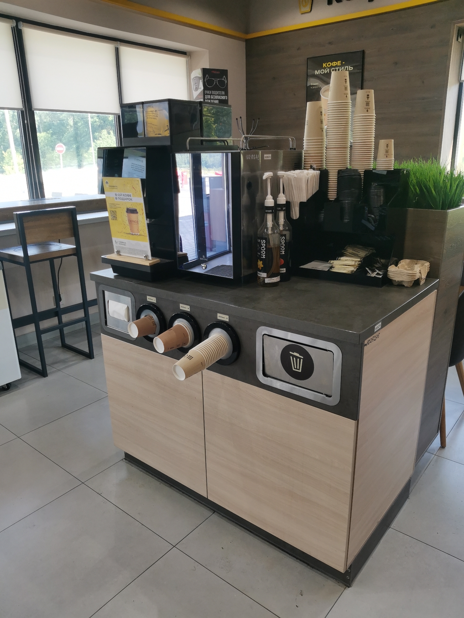 This is a photograph of a coffee station in what appears to be a public establishment, possibly a cafe or convenience store, with a focus on self-service. The scene is well-lit, suggesting it is daytime.
The central object is a complex coffee machine with several dispensers and a large stack of paper cups. There are two bottles of syrups, one labeled "SPOOM" and the other "AMEL," suggesting flavor customization options. Next to the syrups are packets of sugar and creamer, and some small, folded cardboard cup holders.
In front of the coffee machine, there is a counter with three openings, each holding a paper cup, ready for use. One cup is light brown, and the other two are beige. Below these openings, the station has wooden cabinet doors. To the left of the coffee machine, there's a high stool at a counter.
On the wall behind the coffee station, there are two signs. One is a poster with the text "КОФЕ - МОЙ СТИЛЬ" (COFFEE - MY STYLE) and a slogan "Бодрит в дороге" (Invigorates on the road). Another sign, partially visible, has text that translates to "DRIVER'S GLASSES FOR SAFE DRIVING."
To the right of the coffee machine, there's a black planter filled with artificial green grass. A stainless steel bin with a trash can symbol is integrated into the counter. The floor is tiled in a light gray color. The overall impression is a clean, modern, and functional coffee service area.