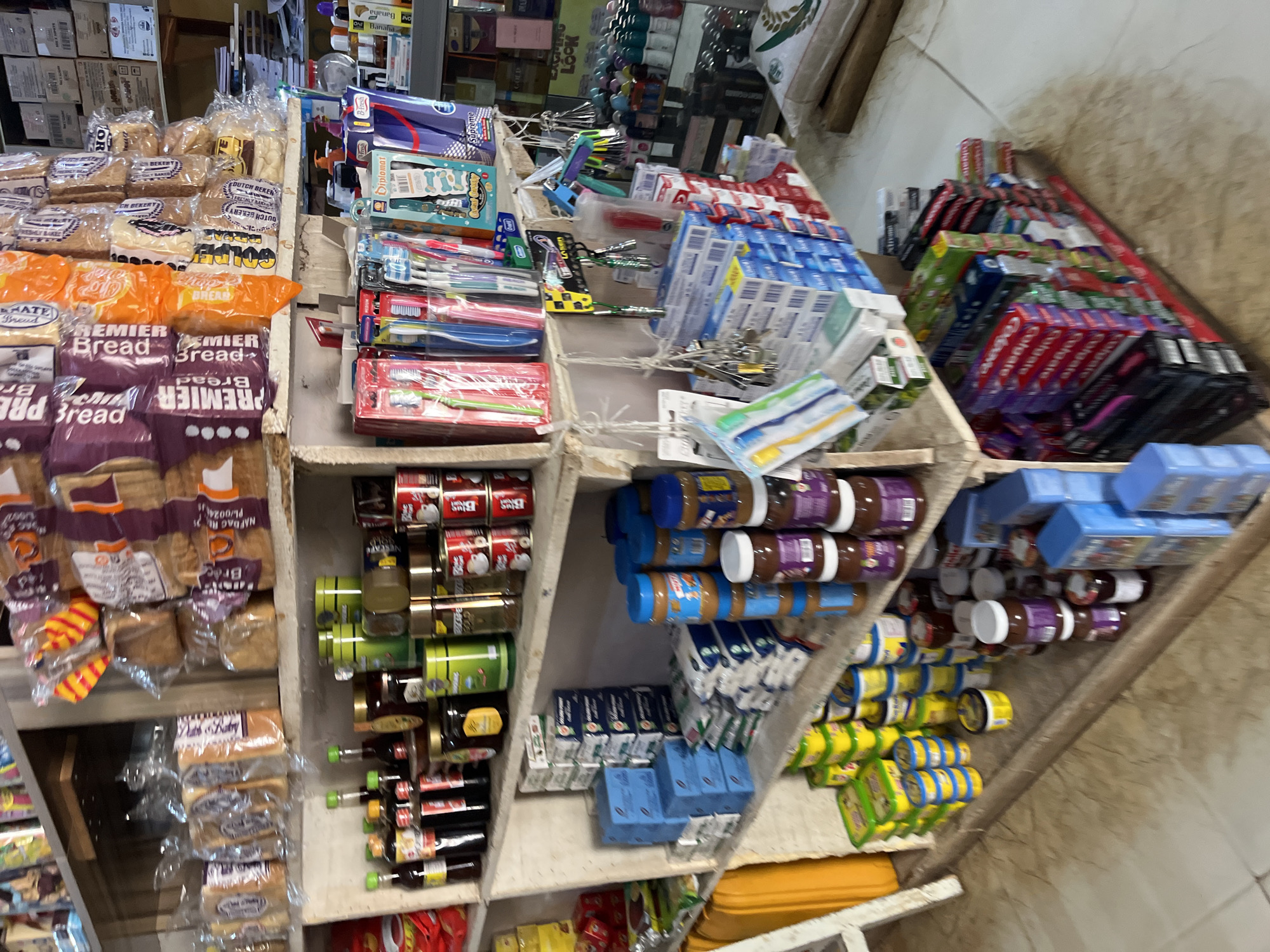 This is a medium shot of a store shelf display in Bukuru, Nigeria, filled with a variety of consumer goods. The shelves are made of rough, light-colored wood and are stacked high with items.
On the top shelf, there are packages of bread in clear plastic wrap, some with purple and orange branding. There are also some smaller items in clear bags and boxes, possibly snacks or baked goods.
The second shelf from the top features an assortment of bottled liquids, including what appear to be sauces or condiments in dark bottles with colored caps (green, yellow, red). There are also jars of jams or preserves, and several round, green cans that might contain food products. Smaller cans with red and white labels are also visible.
The third shelf is dominated by stacks of blue cardboard boxes. Interspersed with these are some white, bagged products. There are also several jars of peanut butter or similar spreads with blue and white labels, stacked vertically.
The bottom shelf is a mix of items. There are more jars of spreads with purple labels, and smaller jars of what appear to be jams or preserves. To the left, there are stacks of yellow containers. Further to the right, on a lower level, are rows of packaged toothbrushes in clear plastic, some with colored handles and bristles. There are also boxes of toothpaste, with prominent red and white "Colgate" branding, and other brands in blue and green packaging. Some loose toothbrushes are also visible.
The overall impression is of a retail environment with a diverse range of products, indicating a general store or a section of a larger market. The lighting is somewhat dim, but the items are clearly visible. The floor appears to be tiled with a light-colored, mottled pattern.