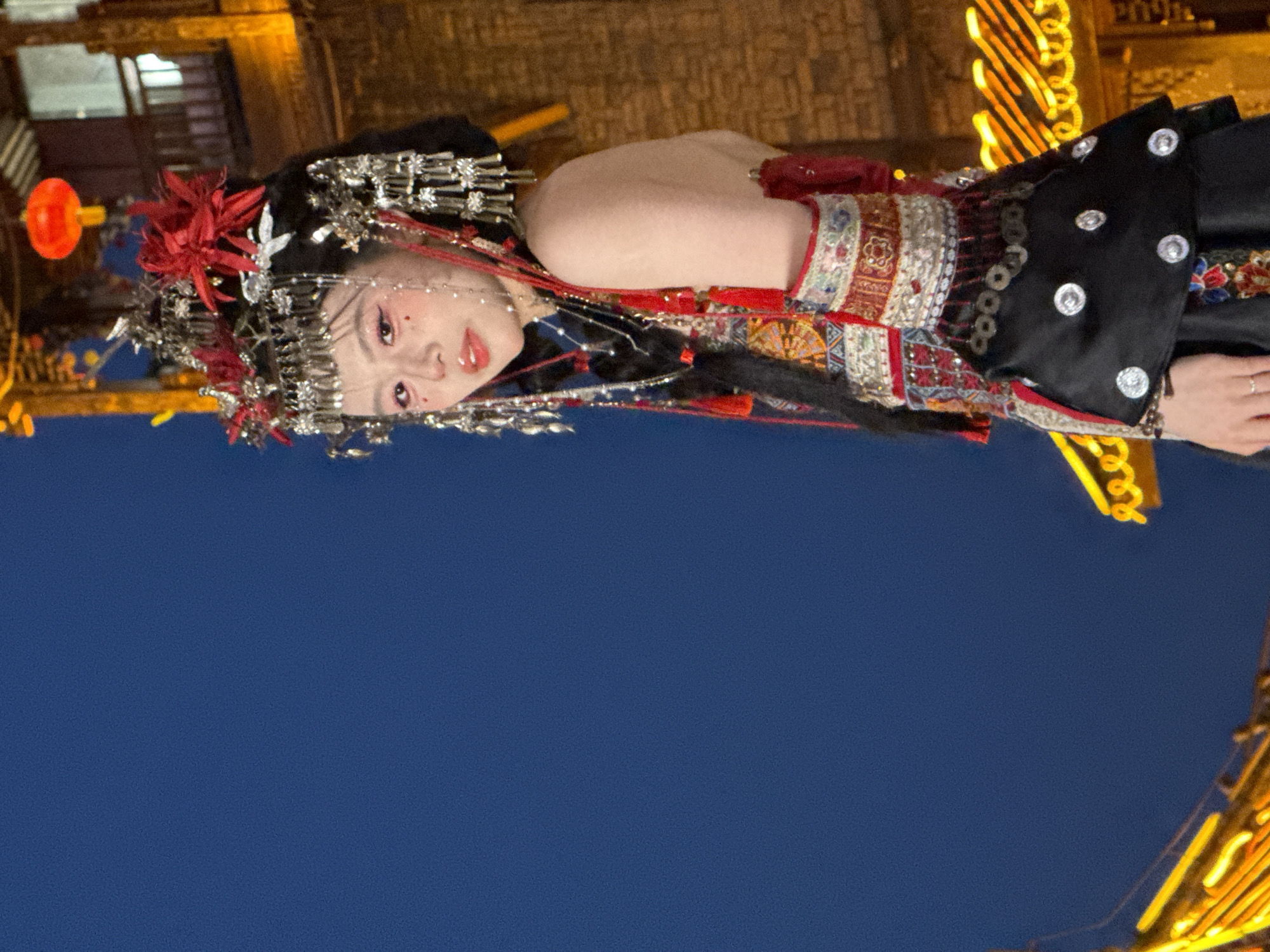 A person in traditional Chinese attire is photographed against a clear blue background and partially visible urban background. The person is wearing a headpiece with red and silver decorations and a black garment adorned with silver circular designs. The Yuzhong District in China is alluded to by visible buildings and streetlights.