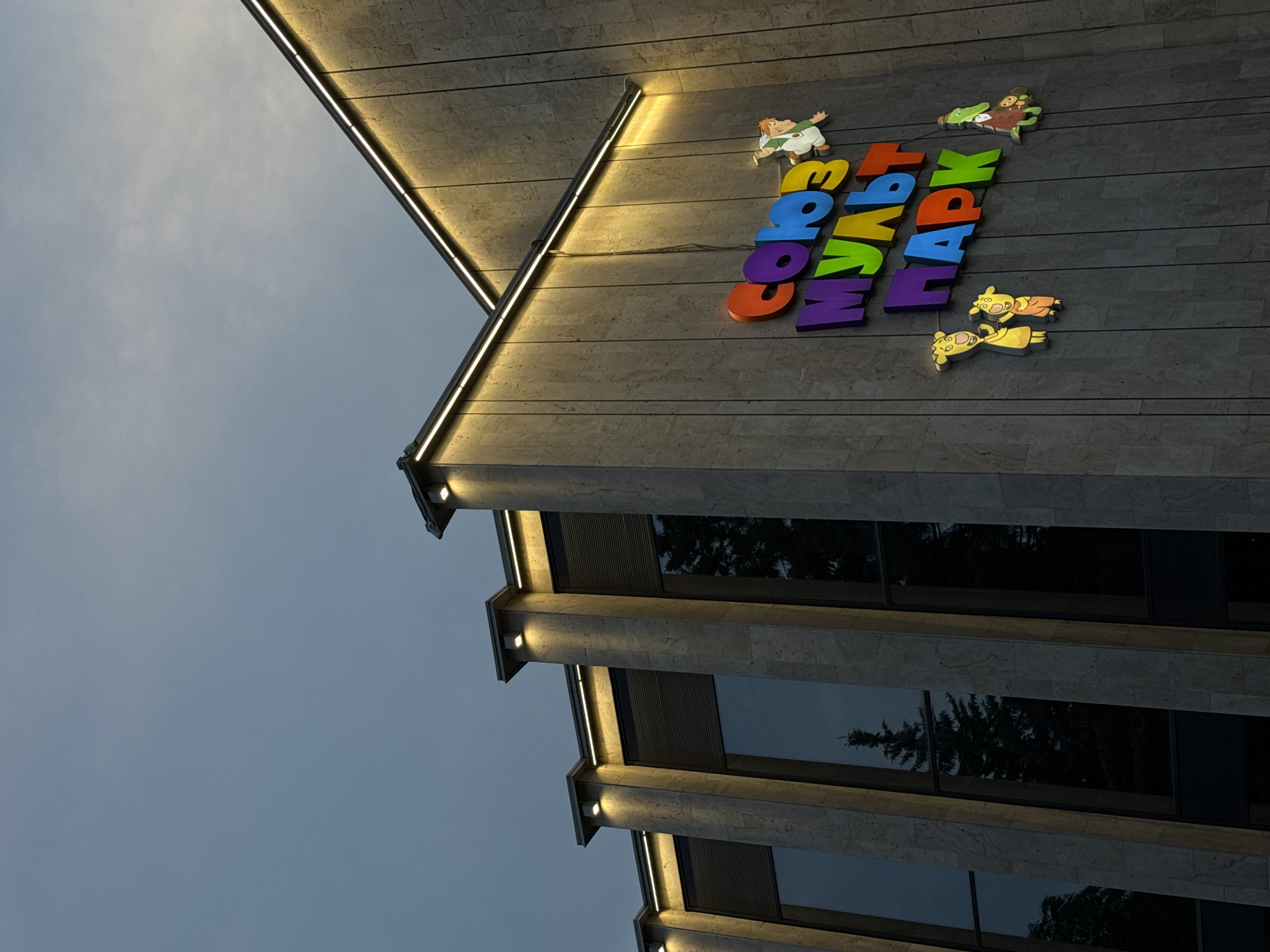 The image shows the exterior of the "Soyuzmultpark" building in Khimki, Russia, under a cloudy sky. The building features a modern architectural design with stone cladding and large windows. Above the windows, a row of lights illuminates the facade. A prominent sign with colorful, three-dimensional letters spells out "СОЮЗ МУЛЬТ ПАРК" (Soyuz Mult Park). Small cartoon character cutouts are attached to the wall near the sign. The windows reflect the surrounding trees and the sky. The overall scene suggests a public entertainment venue.