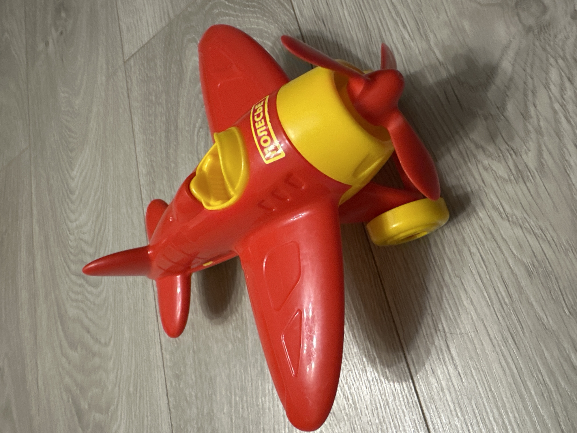 A red and yellow plastic toy airplane rests on a gray wooden floor. The airplane is angled with its nose to the right and its tail to the left. The body of the plane is red, with a yellow cockpit area and yellow accents on the front. A red propeller is attached to the front. The wings are red, and the tail fin is also red. There is a yellow label on the side of the plane with black lettering that appears to be Cyrillic. The floor has a wood grain pattern. There are no people or other objects visible in the image. The scene is indoors and appears to be a residential setting. The location is identified as Energetik, Russia.