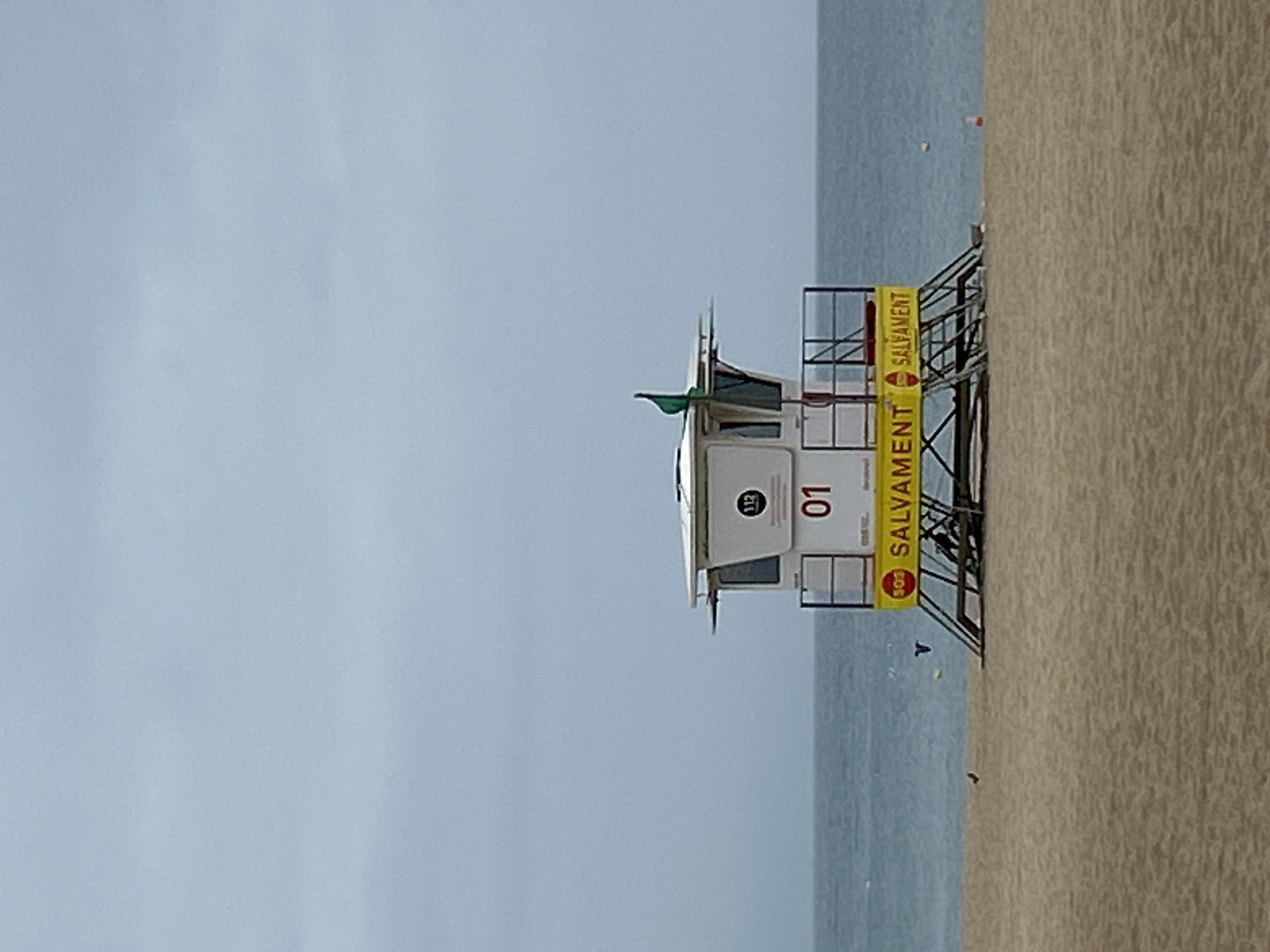 This image captures a tranquil beach scene dominated by a lifeguard tower under an overcast sky. The scene is oriented vertically, with the sky occupying the upper left, the sea in the middle, and the sandy beach on the right.
The central subject is a white, multi-level lifeguard tower, prominently featuring the number "01" on its observation cabin. A small green flag flies from the top of the cabin. The tower stands on a metal framework, adorned with bright yellow banners displaying the word "SALVAMENT" in black capital letters, indicating "lifesaving" or "rescue" in Catalan. One banner clearly shows "SOS SALVAMENT". A circular emblem with the emergency number "112" is also visible on the tower.
To the left of the tower is the calm, muted blue-grey sea, reflecting the light from the overcast sky. Several small buoys are scattered across the water. The sandy beach, with its light brown color and visible texture, occupies the right side of the frame, extending from the foreground towards the waterline.
The sky is a uniform, pale blue-grey, suggesting an overcast day. No people are visible, lending a quiet and serene atmosphere to the scene. The presence of the lifeguard tower and the Catalan text "SALVAMENT" are strong indicators of a beach location, consistent with Barcelona, Spain.