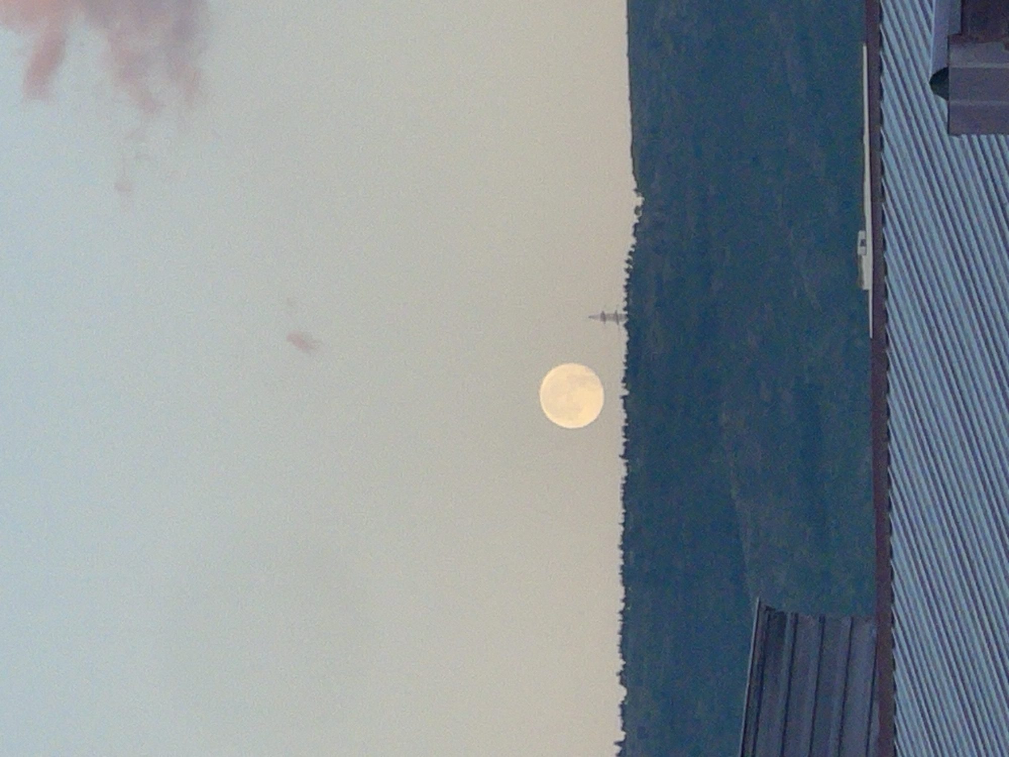An image captured at dusk or dawn in Tbilisi, Georgia, shows a large, full moon positioned centrally above a dark, tree-covered hillside. On the crest of the hill, a slender tower with a cross-like structure at its peak is visible. The foreground features parts of two roofs: the upper one has horizontal wooden siding, and the lower one is covered in corrugated metal. The sky is a pale gradient of blue and grey, with a hint of pink cloud in the upper right corner. The overall scene is serene, with the moon being the dominant luminous element against the darkening landscape.