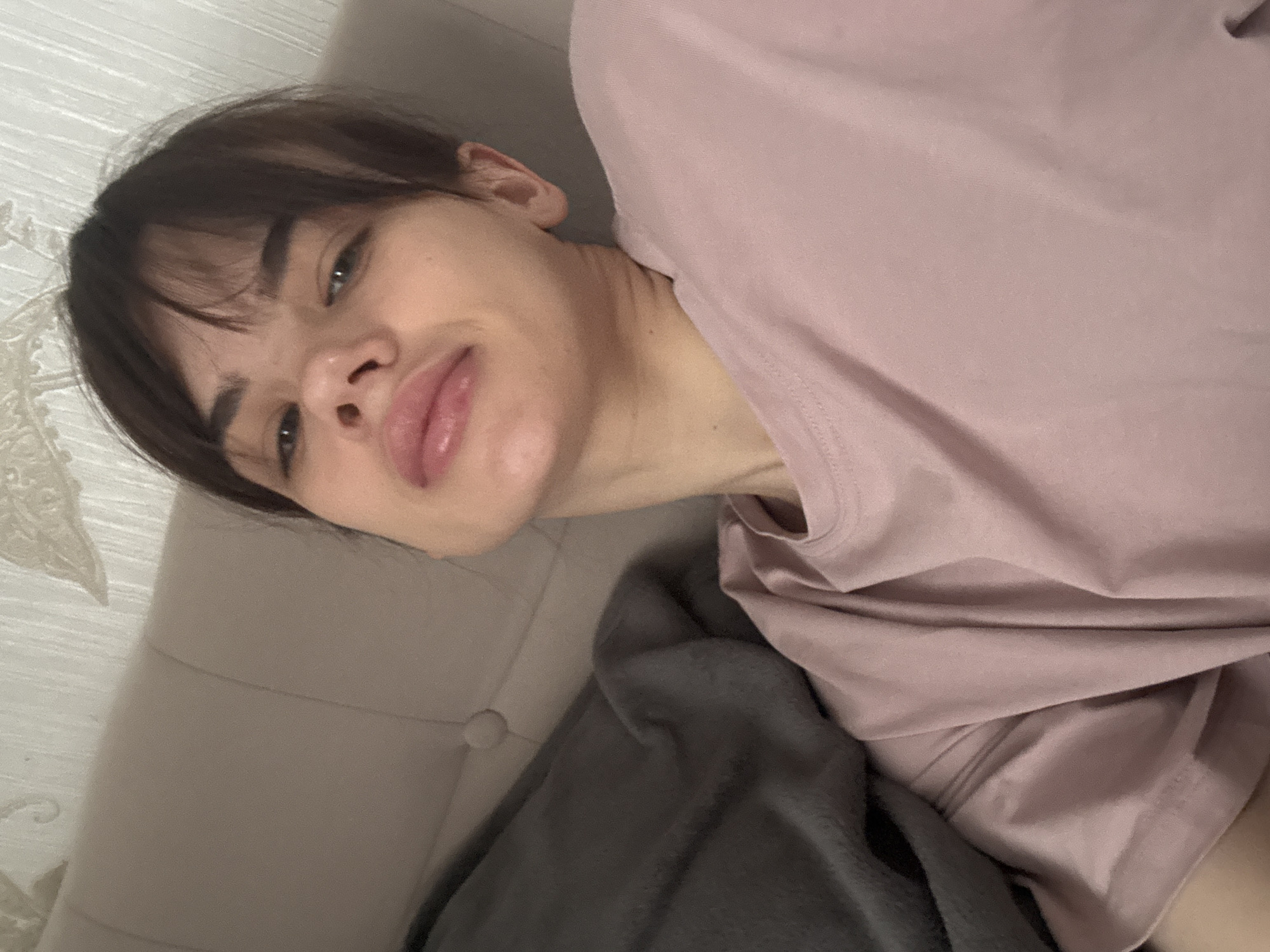 A woman with short, dark hair and a fringe is lying on a light-colored tufted sofa, possibly a chaise lounge, covered with a dark grey blanket. She is wearing a pale pink t-shirt. The location is unknown.