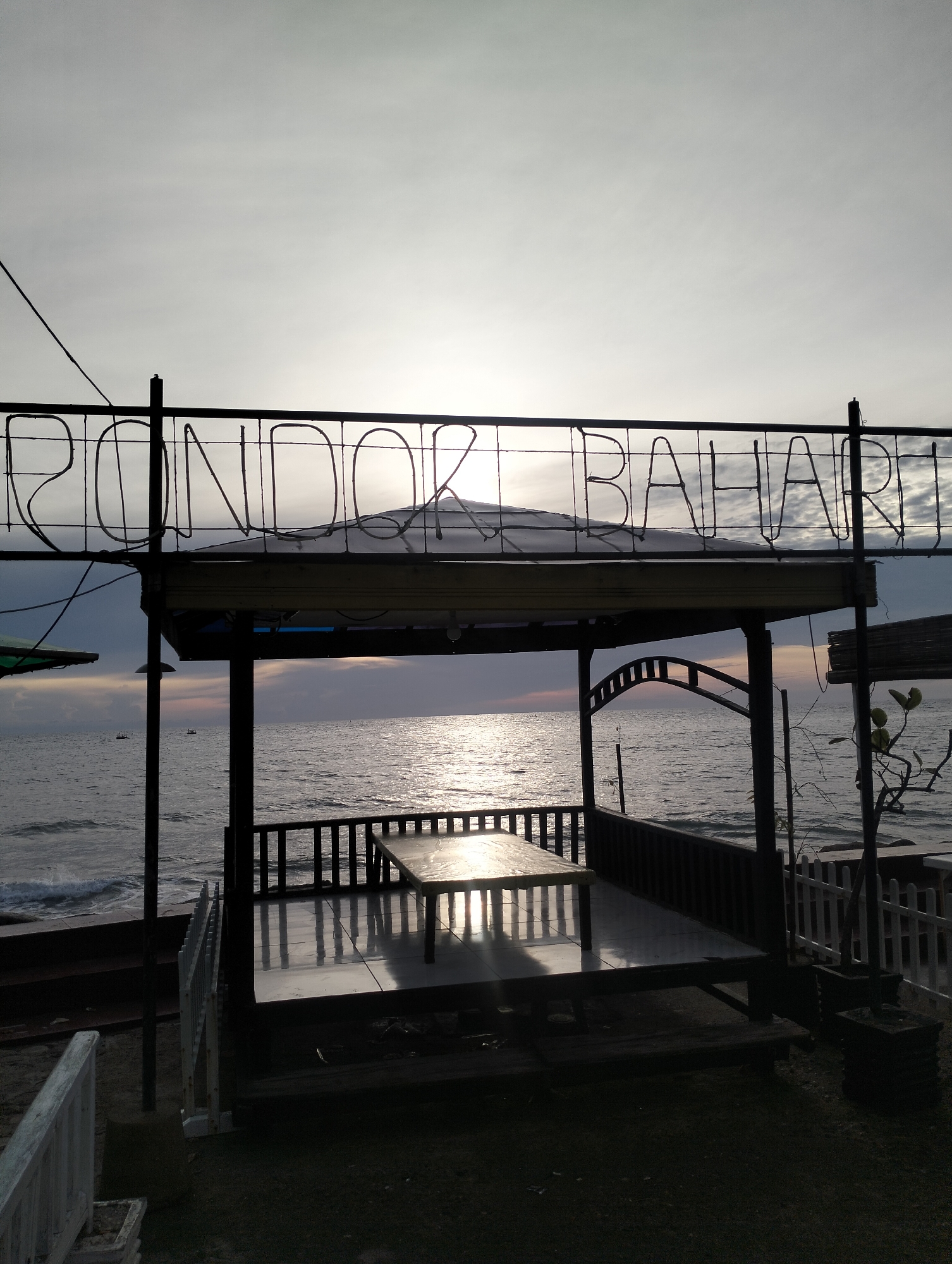 The image presents a serene coastal scene in Lhokseumawe, Indonesia, captured during either sunrise or sunset. A prominent open-air gazebo or shelter, featuring a simple wooden table and benches, overlooks the calm ocean. The structure is largely silhouetted against the bright, partially cloudy sky and the sun's reflection on the water.
Above the gazebo, a large sign crafted from bent metal tubing spells out "RONDOR BAHARI." The low sun casts a strong, sparkling path of light across the ocean's surface, which also illuminates the tabletop within the shelter, creating a striking reflection. Distant clouds appear darker near the horizon over the sea, while the sky directly above is brighter. Two small boats are visible in the distance on the water. To the right of the gazebo, a white picket fence marks a boundary, and a small plant or tree stands nearby. The scene conveys a sense of tranquility and quiet contemplation by the seaside.