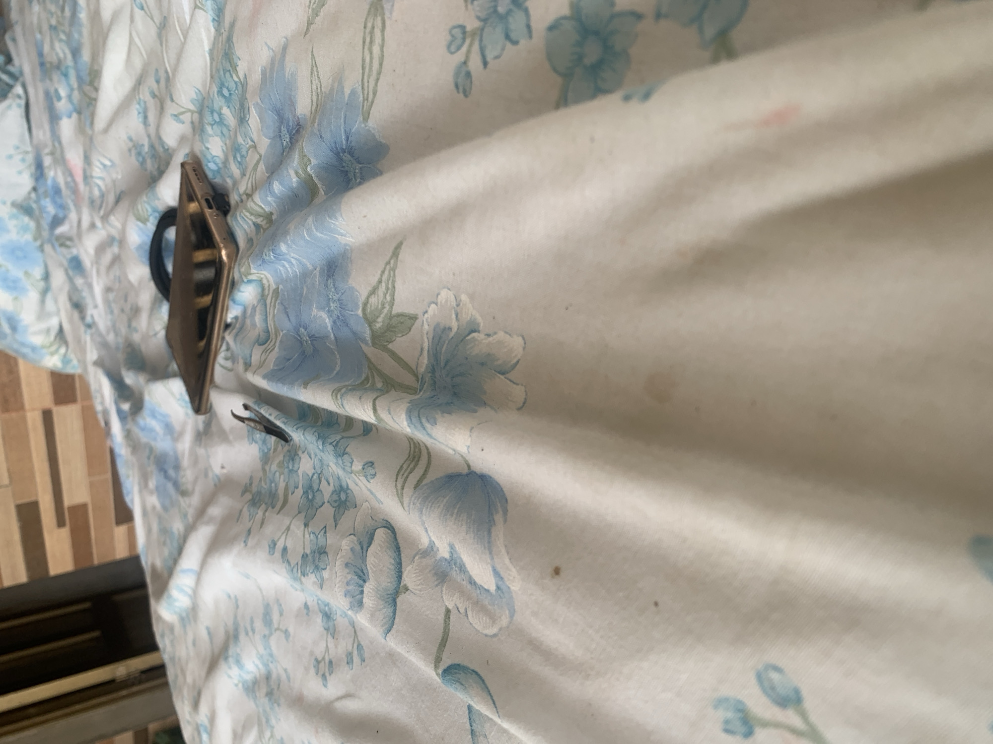 An indoor scene depicts a bed covered with a white sheet patterned with light blue flowers and green leaves. The sheet is rumpled and exhibits multiple creases. A gold-colored smartphone lies face down on the sheet, with its USB-C port visible, and a black loop-shaped object or cable is positioned near it. To the left of the phone, a pair of small, dark metallic tweezers rests on the sheet. Several small, irregular brown stains and one faint pink stain are visible on the white fabric of the sheet. In the background, a wall constructed of horizontal rectangular tiles in varying shades of brown and beige is partially visible, alongside a dark vertical element, possibly a door jamb or furniture. The specific city of Santa Ana, Venezuela, cannot be determined from the visual information present in the image. No individuals are present. The scene is static.