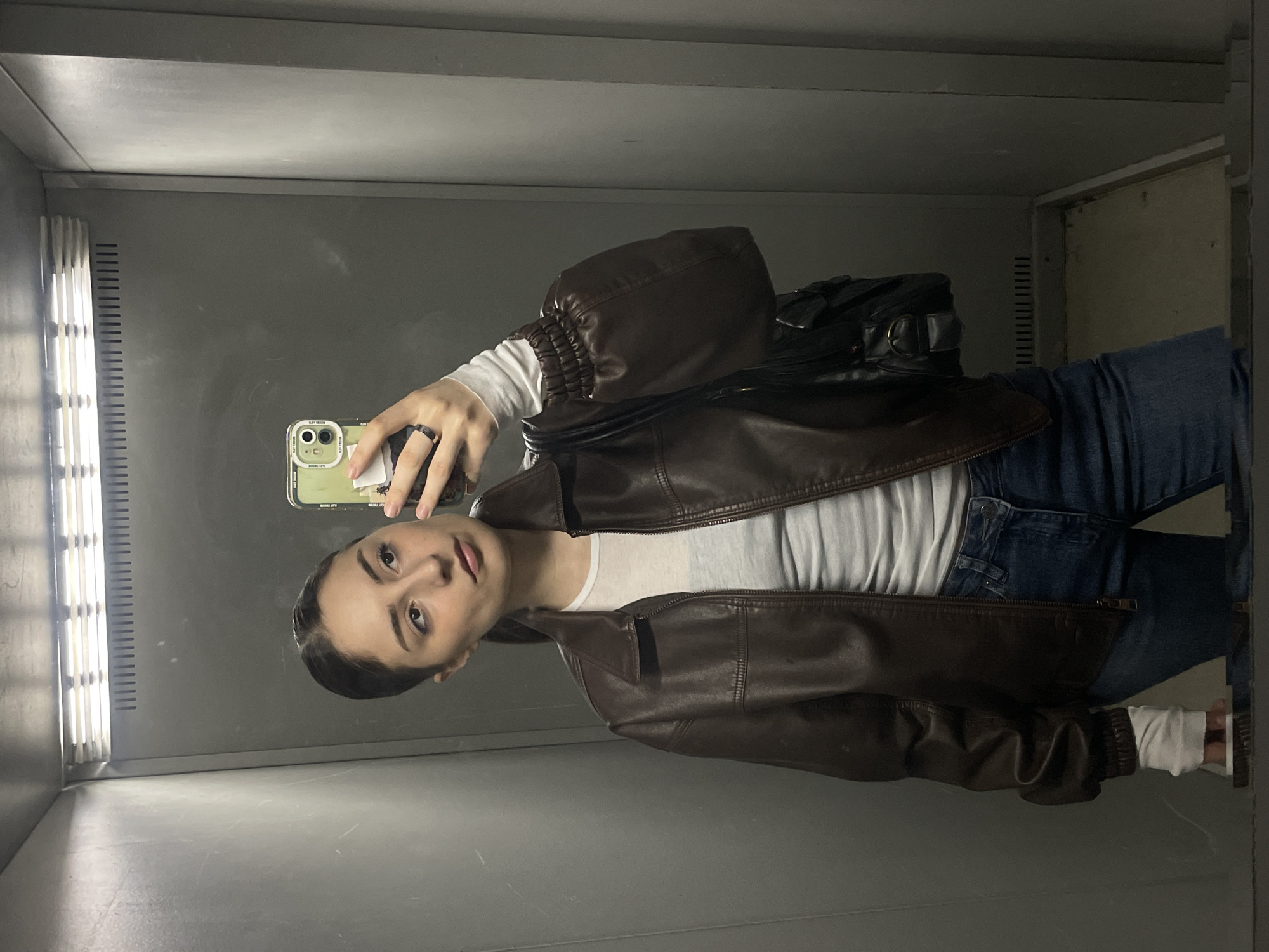 A young woman is taking a mirror selfie in what appears to be an elevator with gray walls and a fluorescent light. She is wearing a brown leather jacket over a white long-sleeved shirt and blue jeans. She is holding a smartphone to take the picture. There is a black bag on her shoulder. The location cannot be confirmed as Moscow, Russia from visual elements alone.