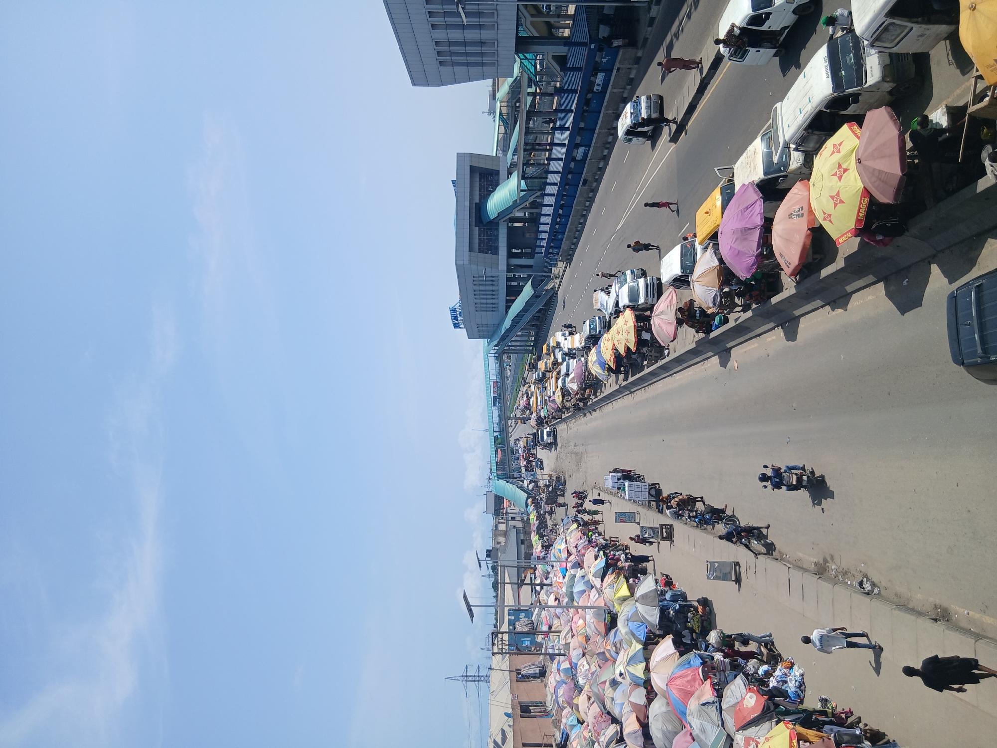 The image depicts a busy street scene in Alaiyabiagba, Nigeria. A wide road runs through the center, with rows of vendors lining both sides. The vendors are sheltered under colorful umbrellas. Many pedestrians are walking on the sides of the roads. Motorbikes weave through the traffic on the road. A large building with a pedestrian bridge spans the road in the background. The sky is clear and blue.