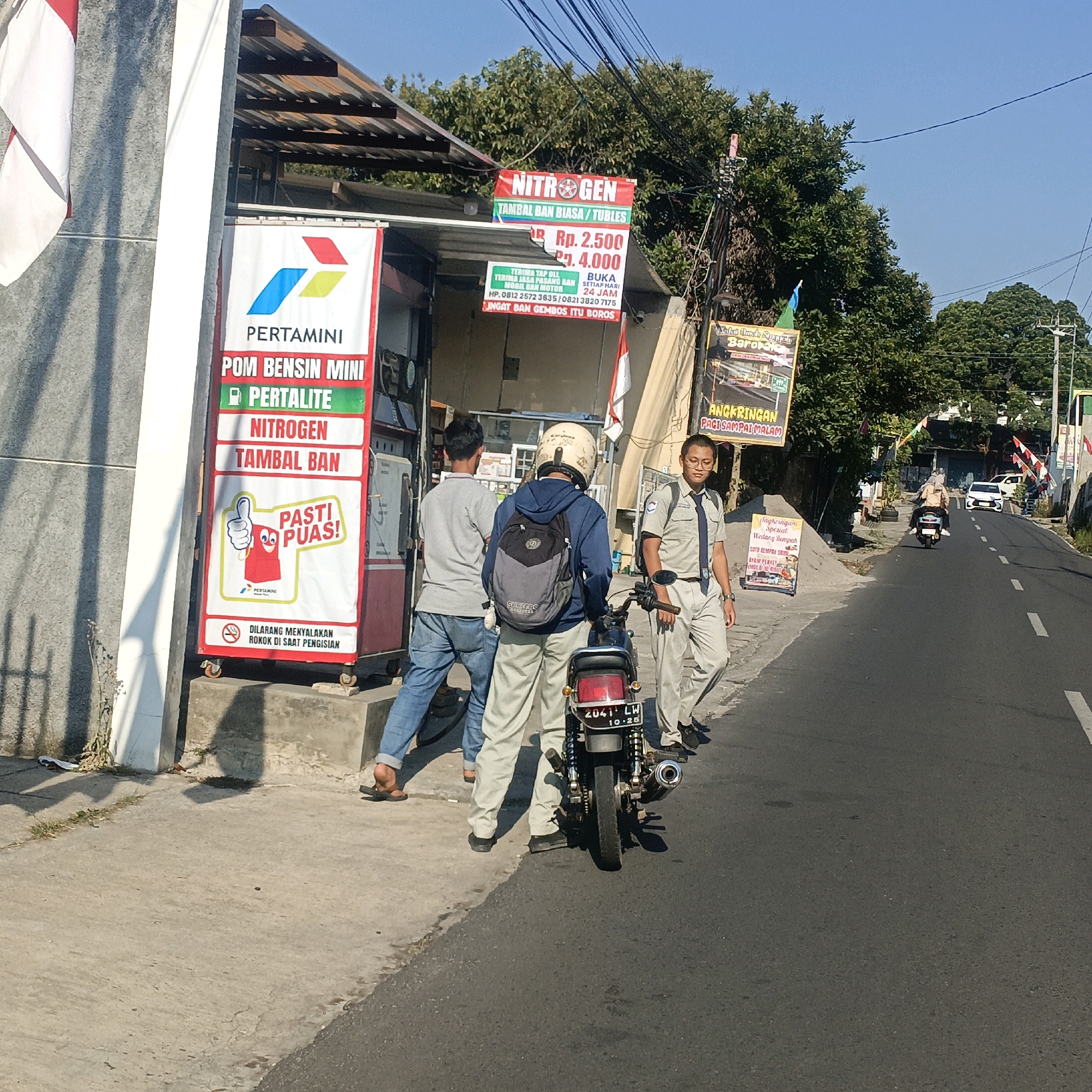 This is a street-level photograph taken outdoors in Salatiga, Indonesia, on a sunny day. The scene depicts a small gas station and convenience store, likely a Pertamini, located alongside a paved road.
In the foreground, to the left, is a white and grey striped pillar supporting an awning. A large red and white banner for "PERTAMINI" is prominently displayed, advertising "POM BENSIN MINI" (Mini Gas Station), "PERTALITE" (a type of gasoline), "NITROGEN," and "TAMBAL BAN" (Tire Patching). The banner features a cartoon thumbs-up character and the text "PASTI PUAS!" (Definitely Satisfied!). Below this is a warning sign stating "DILARANG MENYALAKAN ROKOK DI SAAT PENGISIAN" (Forbidden to light cigarettes during filling).
Three individuals are present at the gas station. One person, wearing a grey t-shirt and blue jeans, stands on the sidewalk, partially obscured. Another person, wearing a blue hooded jacket and a light-colored helmet, is seated on a dark-colored motorcycle, facing away from the camera. The motorcycle has a license plate that reads "2041 LW 10-25". A third person, dressed in a light-colored uniform consisting of a collared shirt with a tie and light-colored pants, stands to the right of the motorcycle, facing towards the road and slightly towards the camera.
Behind the gas station, a roof structure is visible, and further back, lush green trees form a dense canopy against a clear blue sky. Overhead, numerous electrical wires are strung across the scene.
The road in the background is asphalt with white lane markings. Several vehicles are on the road: a motorcycle with two occupants is traveling away from the camera, and a white car is visible further down the road. Another smaller sign, possibly advertising food, is positioned near the road on the right side of the image. The overall impression is of a typical roadside service area in an Indonesian city.