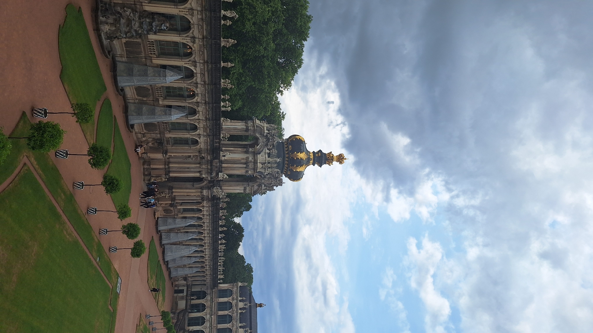 The image depicts the Zwinger palace in Dresden, Germany. The scene is captured from an elevated perspective, looking down upon a courtyard with manicured gardens and a long, arched arcade. In the background, a grand baroque building with a distinctive golden crown-shaped dome, adorned with statues, stands prominently. The sky above is dramatic, with a mix of dark, brooding clouds and patches of bright blue sky.
In the courtyard, several rows of neatly planted trees in patterned pots line the pathways. Some areas of the courtyard are paved with reddish-brown material, while others are covered in vibrant green grass, arranged in geometric garden beds. Along the arcade, several people are visible, appearing to stroll or gather. To the left of the arcade, a large, tiered fountain structure is present. The architecture is ornate, with detailed carvings and classical elements visible on the building facades and the arcade. The overall impression is one of historical grandeur and architectural splendor under a dynamic sky.