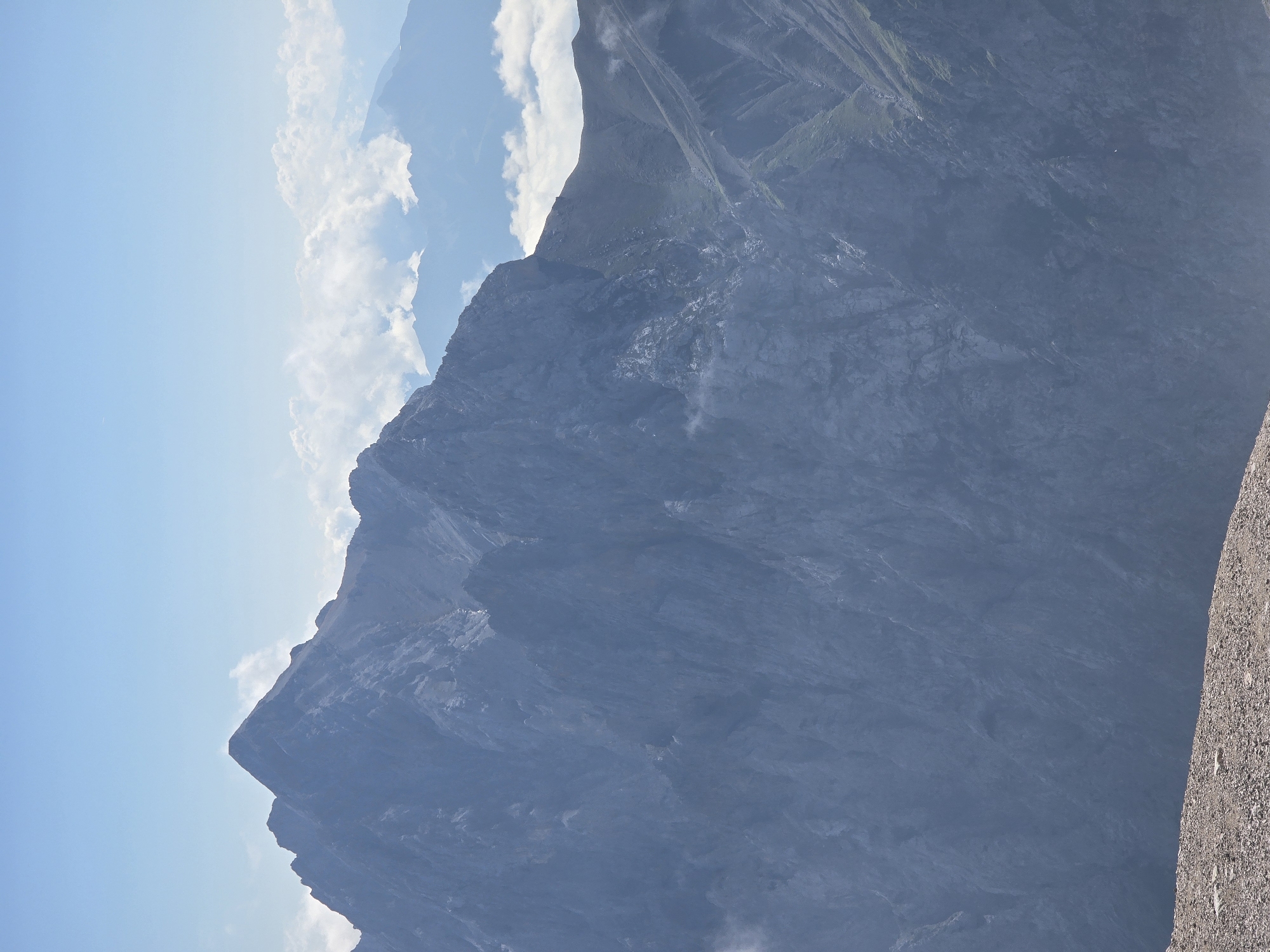 The image depicts a majestic mountain range under a clear blue sky with scattered clouds. The foreground is dominated by a rocky, barren slope. In the mid-ground, a large, dark, craggy mountain peak rises prominently. Its steep slopes are textured with intricate geological formations. Further into the background, other mountain ranges are visible, some shrouded in mist and clouds, creating a sense of depth and distance. The lighting suggests it is daytime, with the sun casting shadows and highlighting the rugged terrain. The scene evokes a sense of natural grandeur and wilderness.
This image is likely of the Zugspitze region near Garmisch-Partenkirchen, Germany, a well-known mountainous area with dramatic peaks and valleys.