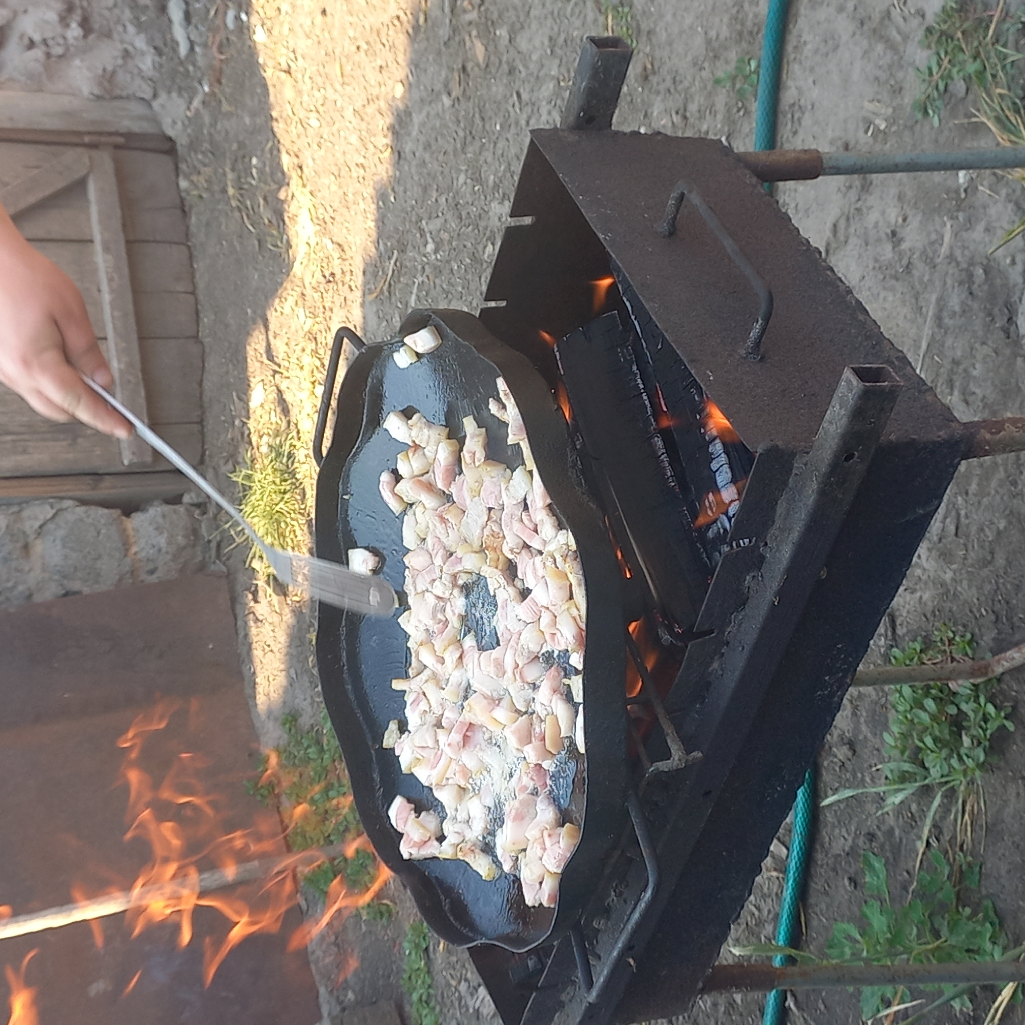 In Klyntsi, Ukraine, a person is seen cooking pieces of pork fat on a large, flat cast-iron skillet placed over an open fire. The skillet is positioned on a rusty metal grill, with burning logs visible beneath it. Flames are actively licking the sides of the skillet, indicating intense heat. The background shows a weathered wooden door and a stone wall, suggesting an outdoor, possibly rural or backyard setting. The ground is covered with dirt and sparse vegetation. A metal spatula is being used to stir the contents of the skillet.