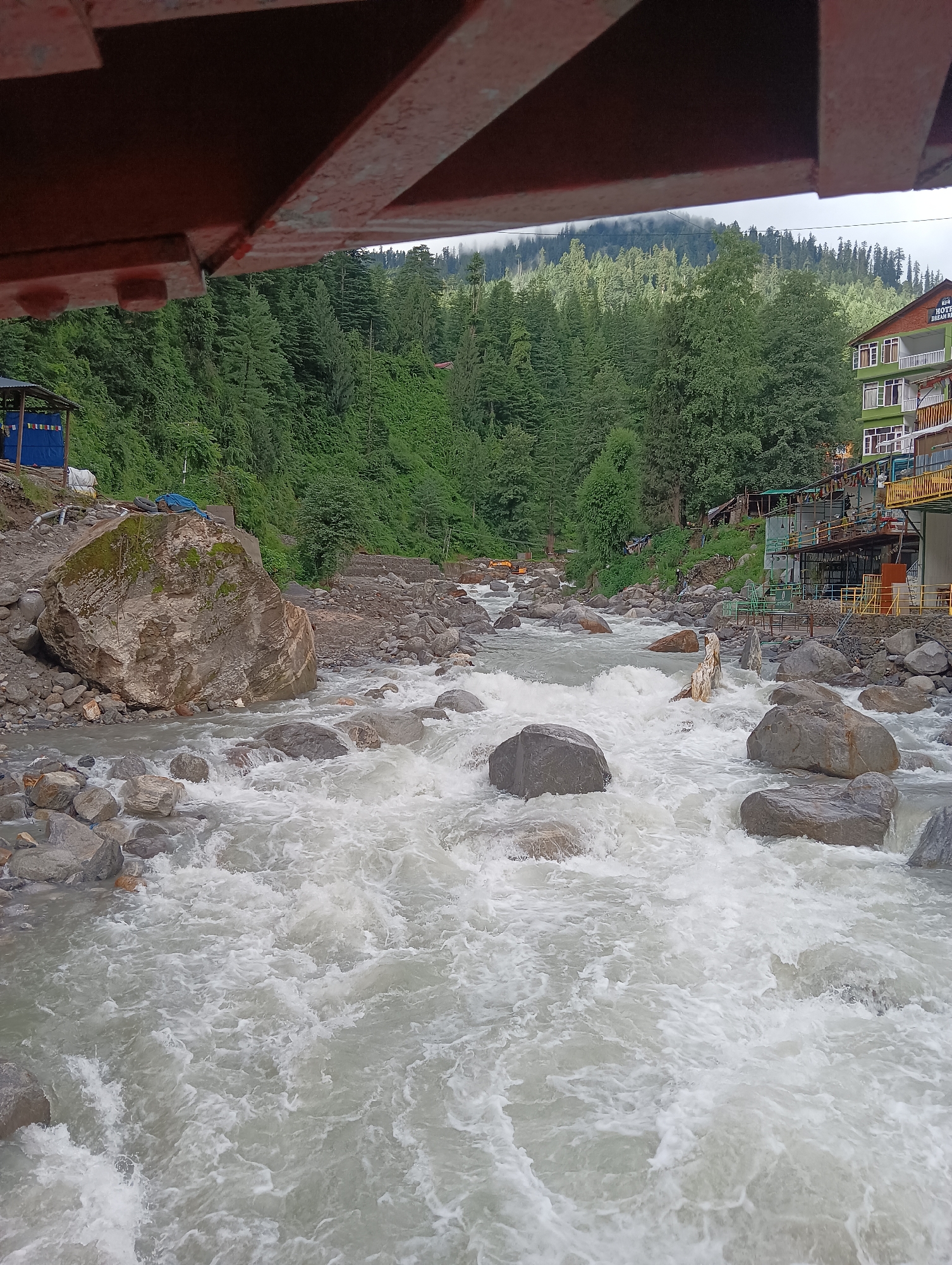 This image depicts a turbulent, milky-white river flowing through a rocky landscape, characteristic of the Beas River in Manali, India. The river is filled with numerous grey and brown rocks of various sizes, some submerged and others exposed. The water is churning with white foam, indicating a strong current.
In the background, a lush green hillside covered with dense pine trees rises steeply. A multi-story building with a green facade and a red tiled roof, identified by signage as "HOTEL DREAM HOME," is visible on the right side of the frame, suggesting it is situated near the riverbank. The architecture of the hotel, with its balconies and windows, appears to be typical of tourist accommodations in the region.
Further in the background, the sky is overcast with clouds, and mist partially obscures the upper reaches of the forested mountains. On the left side of the river, a large, moss-covered boulder is prominently situated near the bank. A small, temporary structure with a blue tarp roof is also present near this boulder, possibly a makeshift shelter or storage area. The overall scene conveys a sense of raw natural beauty and the dynamic power of the river, set against a backdrop of mountainous terrain and a small settlement.