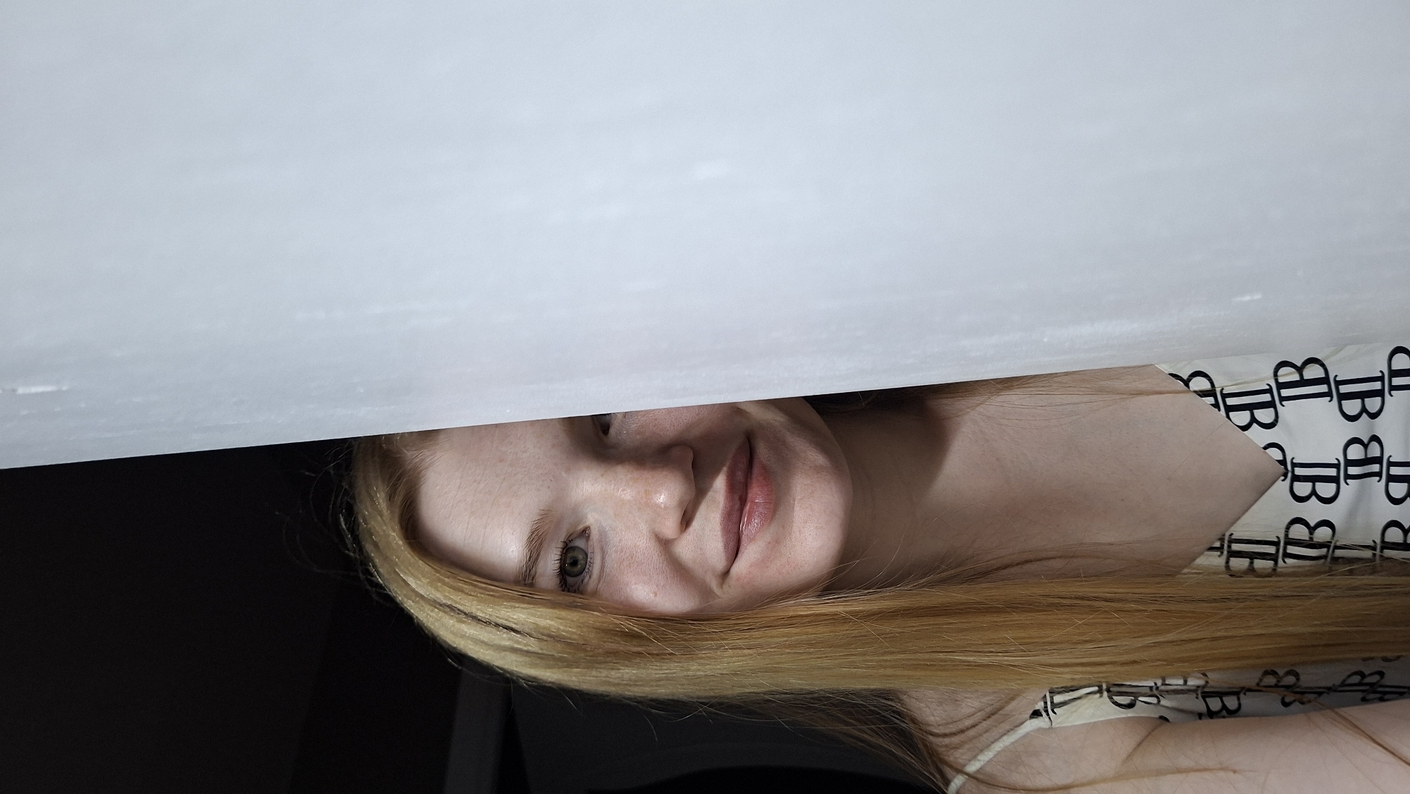 A young woman with long blonde hair and fair skin with freckles is looking at the camera. She is partially obscured by a white curtain on the right side of the image. She is wearing a white top with a black pattern. The background is dark on the left side of the image, suggesting an indoor setting. There is no information in the image to determine its location in the city of Chaikovsky, Russia.