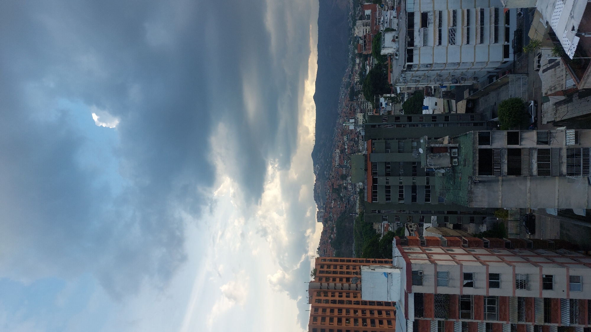 The image shows a view of Caracas, Venezuela. From the vantage point, the viewer sees densely packed buildings, transitioning in size and color from lower in the frame toward the horizon where they meet the backdrop of a mountain range. The architecture is mixed, with a variety of building heights and materials. The sky overhead is partly cloudy, with a mix of dark rain clouds and patches of blue sky. The overall light is soft and diffused, suggesting late afternoon. There are no people visible.