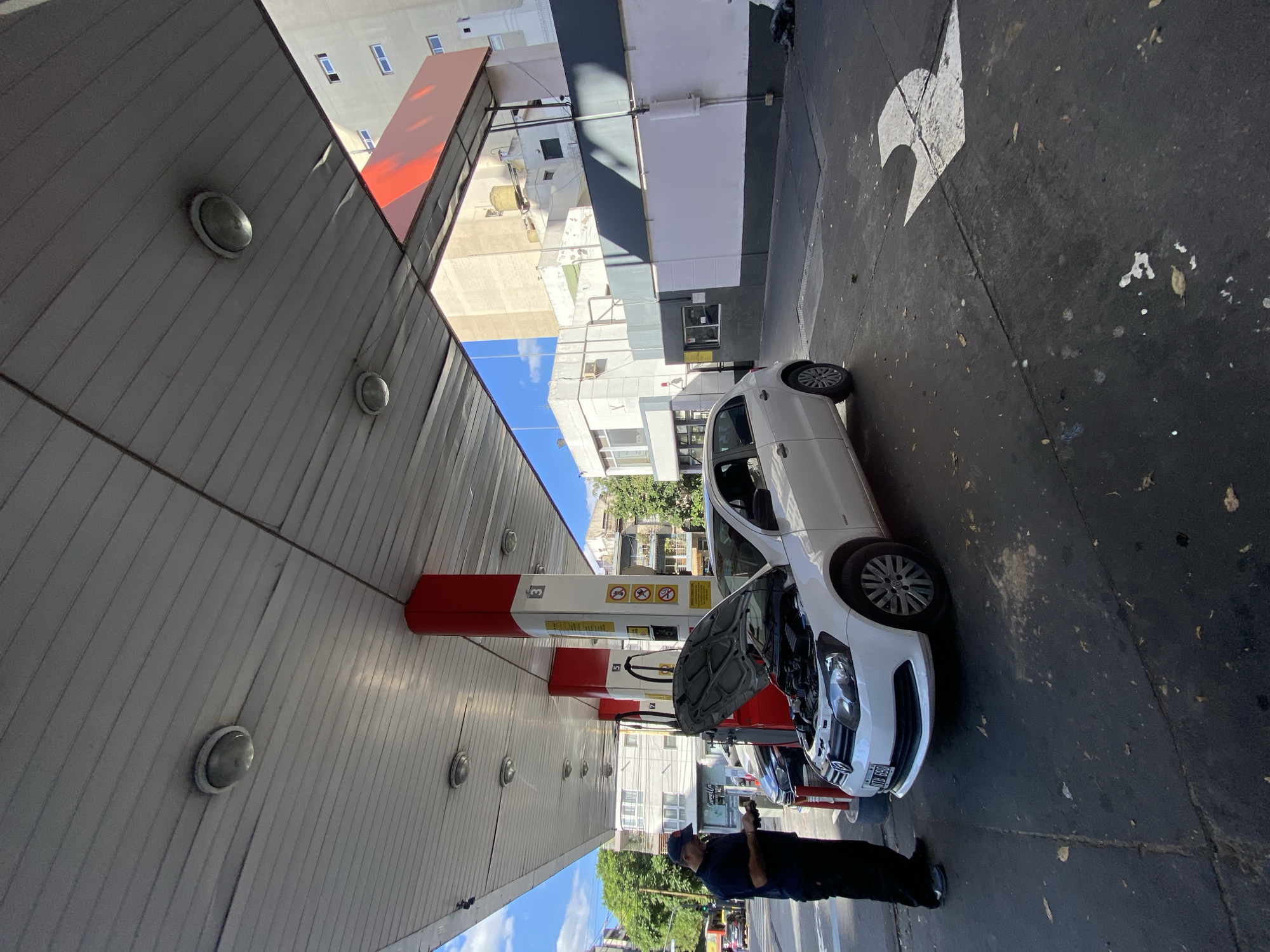 The image shows a white car parked at a gas station in Buenos Aires, Argentina. The car's hood is open, and a person in a dark blue uniform is standing nearby, presumably inspecting or working on the vehicle. The gas station has a red and white canopy, and multiple pumps are visible. The background includes buildings of varying heights and architectural styles, a blue sky with scattered clouds, and street markings on the pavement. The gas station appears to be located on a street with moderate vehicular traffic.