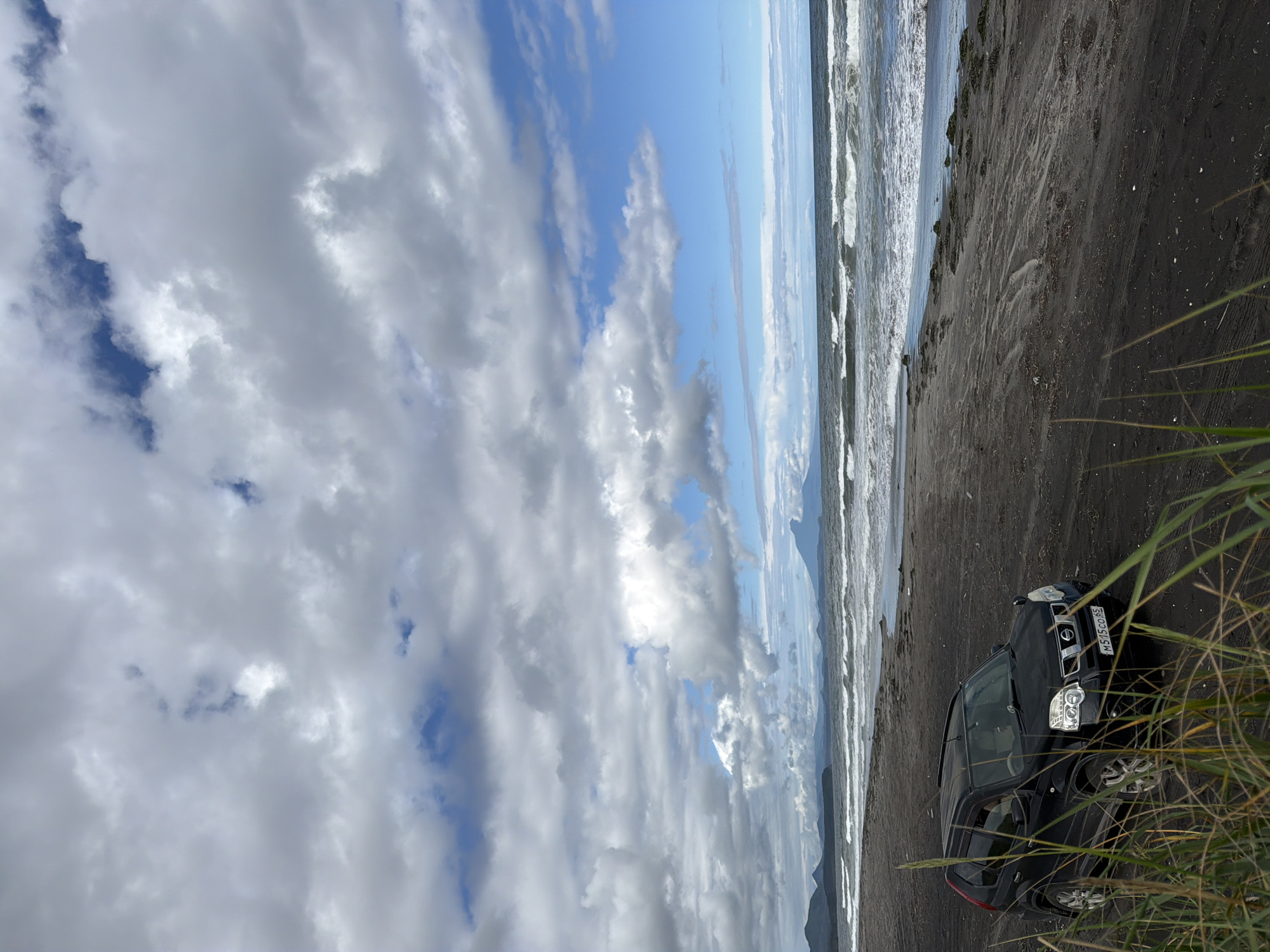 A black Nissan X-Trail SUV is parked on a dark sand beach, facing left. The vehicle has its headlights on. In the background, the ocean stretches to the horizon with waves crashing on the shore. A distant volcano or mountain range is visible on the left, and another, larger volcano is prominent on the right. The sky is partly cloudy with patches of blue. Tall green grass grows in the foreground. The scene appears to be located in Yuzhno-Kurilsk, Russia, likely along the coast. There are no people visible in the image.
