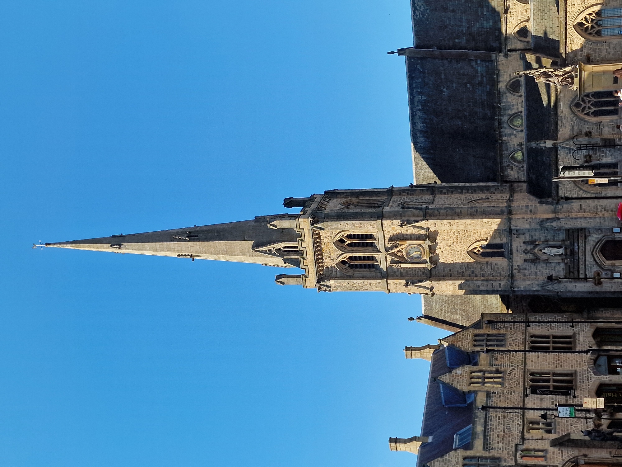 The image captures a striking view of a majestic stone church or cathedral, characterized by its tall, slender spire that tapers upwards against a vivid, cloudless blue sky. The main structure is built from rough-hewn stone blocks, showcasing architectural features such as arched windows, buttresses, and decorative elements like gargoyles extending from the roofline. A prominent clock face is visible on the tower, and below it, a niche contains a white statue.
The bright, clear sunlight indicates a daytime scene, casting strong shadows that highlight the textures and depth of the ancient stonework. To the right and below the main church, an adjacent multi-story stone building is visible, featuring multiple windows, gabled roofs, and similar traditional stonework.
No people are visible in the image, and no active events are taking place; the focus is solely on the architecture. Visible text includes part of a sign on the lower building which appears to read "Town Hall," and a smaller green and white sign nearby. The scene is consistent with the historic architecture found in Durham, United Kingdom.