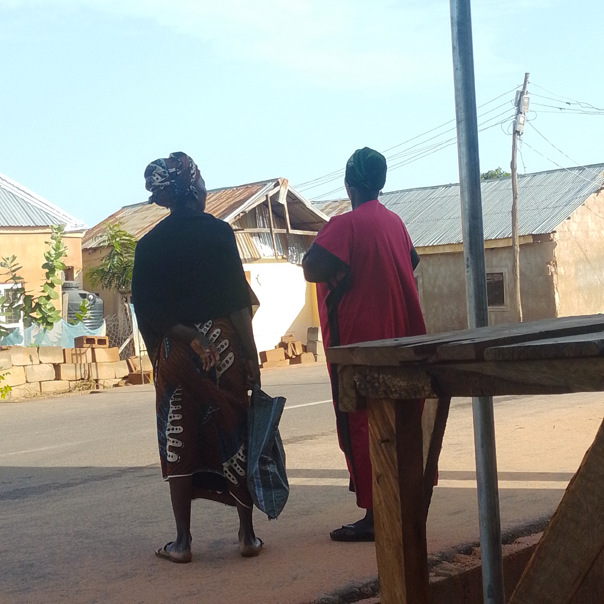 Two individuals stand facing away from the camera on a dusty roadside in Gombe, Nigeria. The person on the left wears a black top and a patterned skirt, with their hands clasped behind their back and a blue bag hanging from their left hand. The person on the right is dressed in a red garment and a green headscarf. In the background, several buildings with corrugated metal roofs are visible, along with a utility pole and overhead wires. The ground is a mix of pavement and dirt.