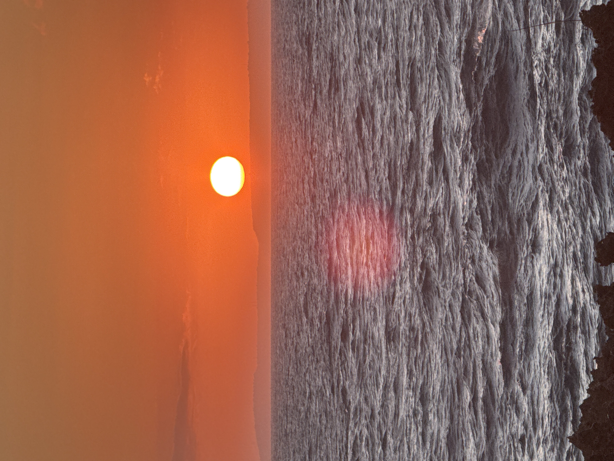 This landscape photograph captures a vibrant sunset over a vast body of water, likely the ocean, with a rugged coastline visible on the far right. The scene is bathed in the warm, diffused light of the setting sun, presenting a striking contrast between the luminous sky and the textured water.
On the left side of the frame, a brilliant, circular sun disc, appearing intensely bright and white-yellow, is positioned low above the perceived horizon. The sky around the sun transitions through a rich spectrum of orange and reddish hues, suggesting the end of the day. The upper left corner shows some subtle, wispy cloud formations, adding slight texture to the otherwise expansive and hazy sky.
The central and right portions of the image are dominated by the water surface, which displays a textured appearance with numerous small, agitated waves or ripples. Despite the fiery sky, the water primarily appears in cooler tones of deep gray and blue. A distinct, elongated reddish glow, clearly a reflection of the sun, shimmers on the water's surface in the mid-right area.
To the far right, a dark, jagged landmass, resembling a cliff or rocky coastline, rises abruptly, forming a strong, dark silhouette against the brighter backdrop of the water and sky. This rugged profile adds depth and an imposing edge to the natural vista.
The overall mood conveyed is serene and majestic, characteristic of a dramatic sunset over a coastal landscape, consistent with locations like Kuta Selatan, Indonesia. No visible text or human activity is present in the image.