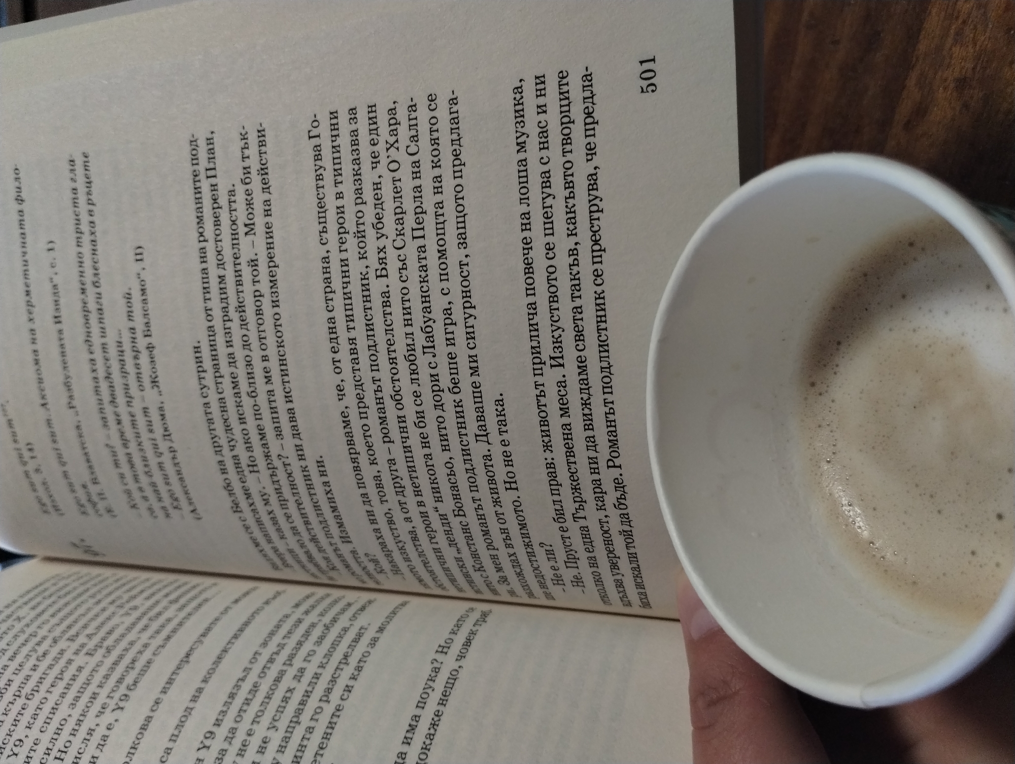 The image captures an intimate indoor scene, likely at a cafe or home in Yambol, Bulgaria, where a person is engaged in reading and enjoying a beverage. The main subjects are an open book and a white paper cup, held by a hand.
The activity taking place is reading, presumably accompanied by sipping coffee or a similar foamy drink. The book is open to page 501, resting on a dark wooden surface. A person's hand with manicured nails holds a white paper cup filled with a light brown, frothy beverage, likely cappuccino or latte.
Notable details include the cozy and relaxed atmosphere suggested by the combination of reading and a warm drink. The lighting appears soft, indicating an indoor setting, but the specific time of day is not discernible.
The visible text in the book is in Bulgarian, with some Latin phrases such as "Ego sum qui sum." The content appears to be a literary discussion, referencing authors like Alexandre Dumas ("Жозеф Балсамо"), and literary characters such as Scarlett O'Hara. Key phrases include "животът прилича повече на лоша музика" (life resembles bad music) and observations about how art shapes perception ("Изкуството се шегува с нас и ни вдъхва увереност, кара ни да виждаме света такъв, какъвто творците биха искали той да бъде"). The text discusses the nature of typical versus atypical heroes in novels and the concept of a "dendі" (dandy), reflecting on the illusion versus reality presented in literature.