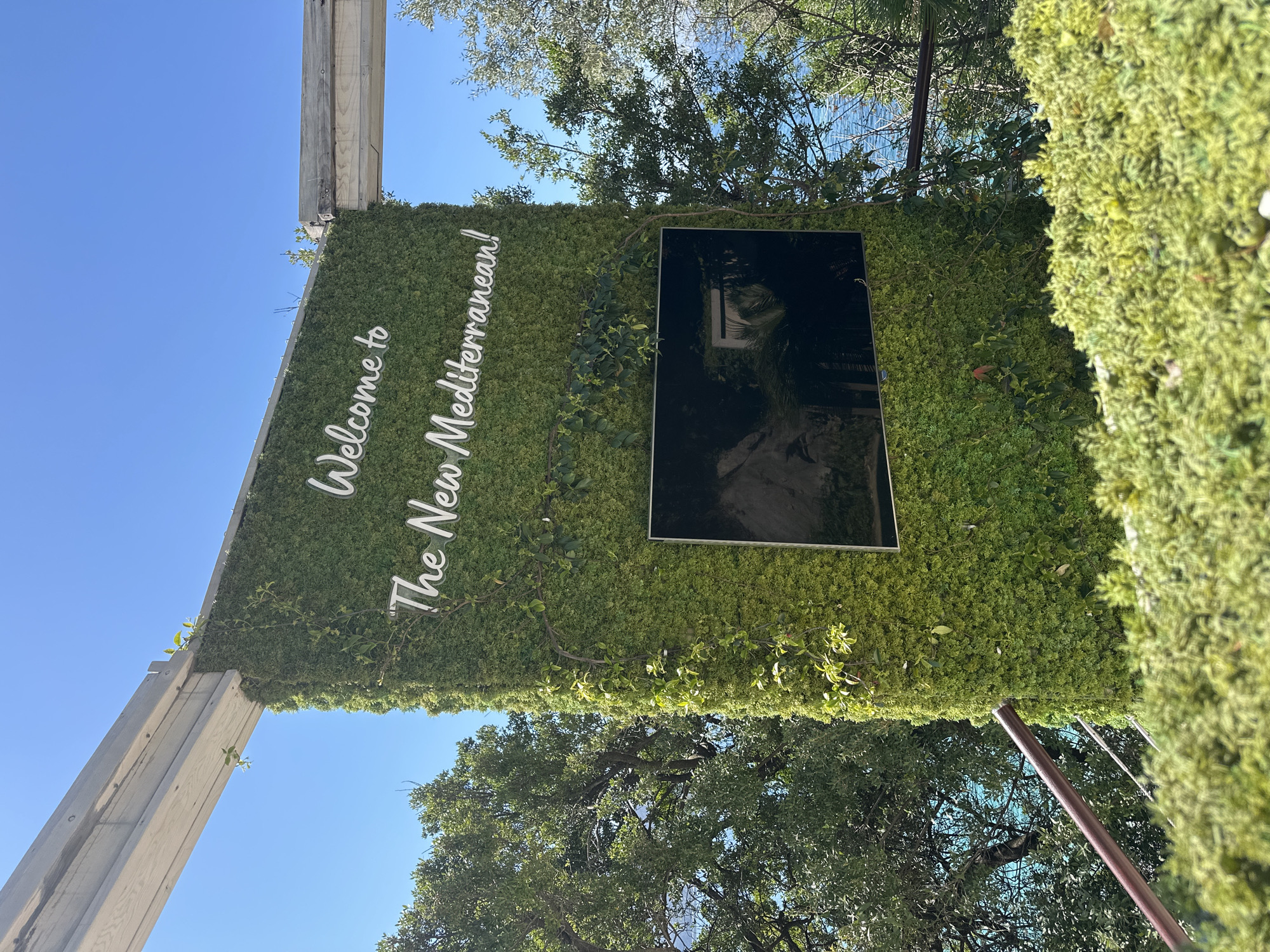 A vertical shot shows a green wall with the text "Welcome to The New Mediterranean!" in white, angled upwards, located in the city of Budva, Montenegro. A black-mirrored rectangular display is inset into the green wall. The green wall is built onto a pale wooden structure and supported by a slim brown pole. Foliage and trees are visible behind and around the wall. The sky is bright blue.
