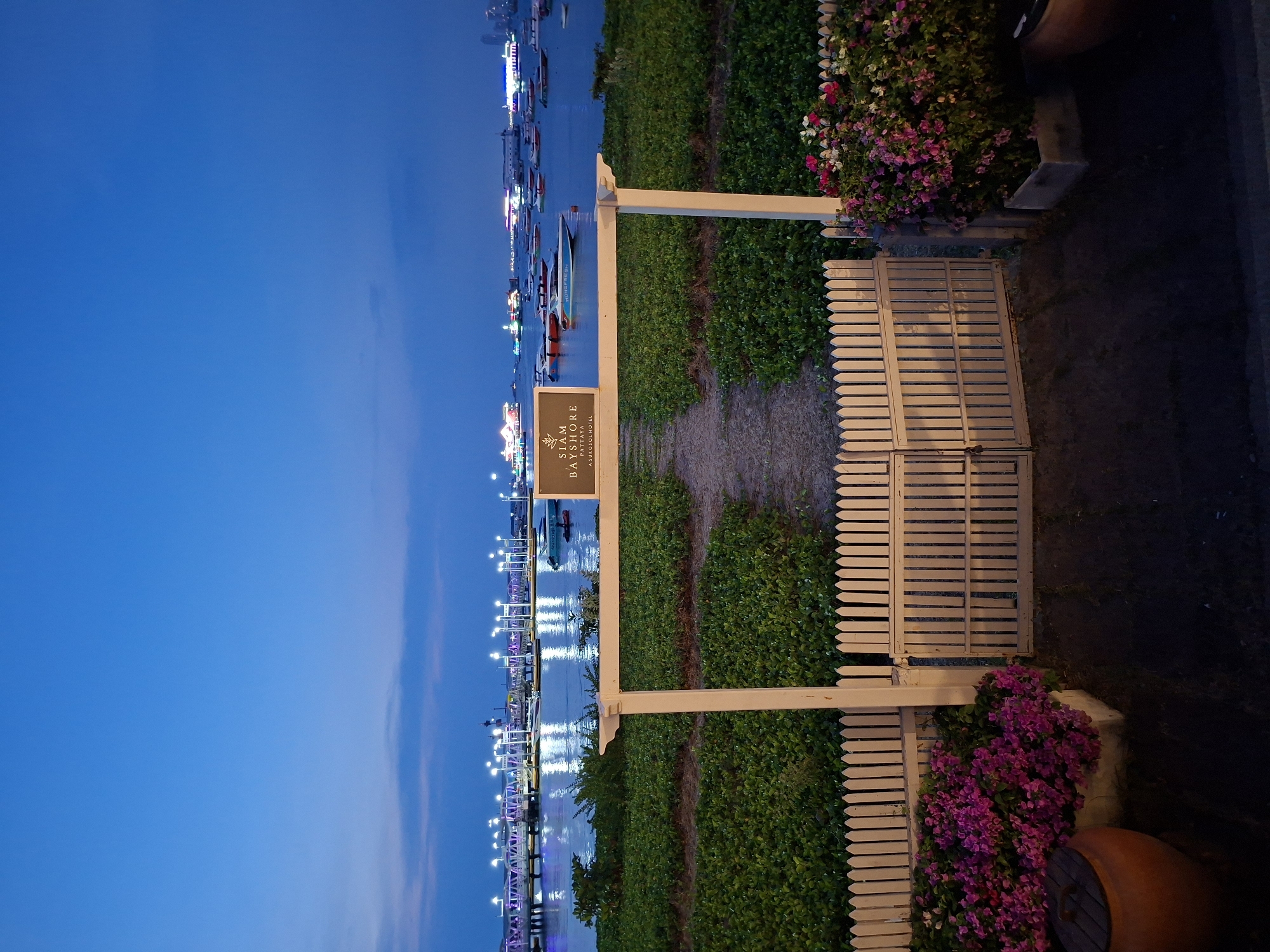 The image depicts a coastal scene in Pattaya City, Thailand, at dusk or night. The "Siam Bayshore Pattaya, A Sukhothai Hotel" sign stands beside a gate, with boats and the pier visible in the water. Plants line the path and adorn the gate area. The setting is a calm waterfront location.