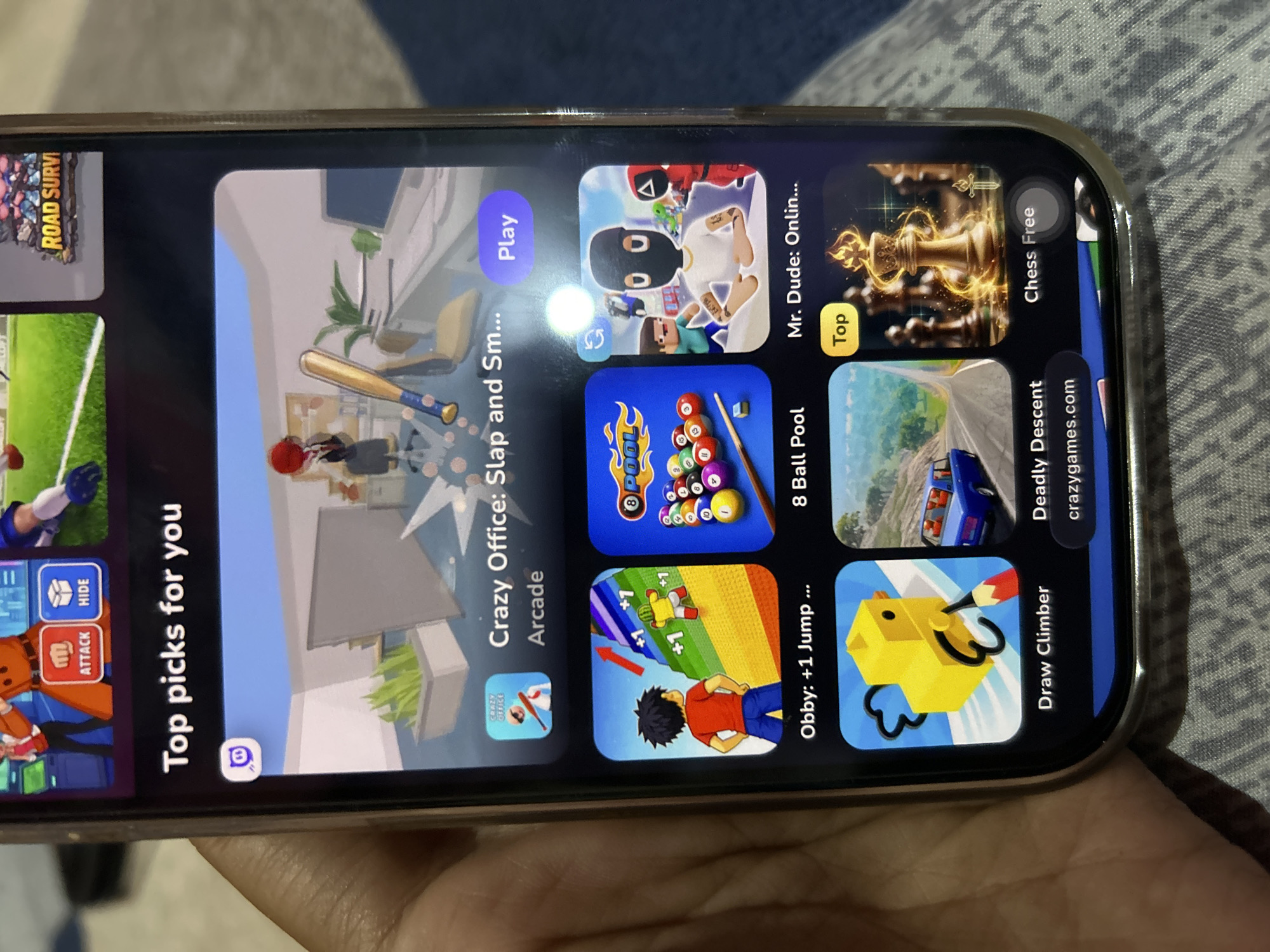 The image is a close-up photograph of a smartphone screen displaying a game launcher or app store. The screen is angled and held by a person's hand, with a patterned fabric visible in the background, likely a bedspread or sofa. The phone's interface shows a section titled "Top picks for you," featuring several game icons and titles.
Prominently displayed are games like "Crazy Office: Slap and Sm...", "Obby: +1 Jump ...", "8 Ball Pool", "Mr. Dude: Onlin...", "Draw Climber", and "Deadly Descent". Some games have "Play" or other action buttons. At the top of the screen, there are smaller icons, one with the Twitch logo and another with game thumbnails, one of which is labeled "ROAD SURVI".
The image does not contain any information or visual cues that would allow for the identification of the city of Depok, Indonesia, or any specific location within it. The content is limited to the smartphone display and the immediate surroundings of the phone.