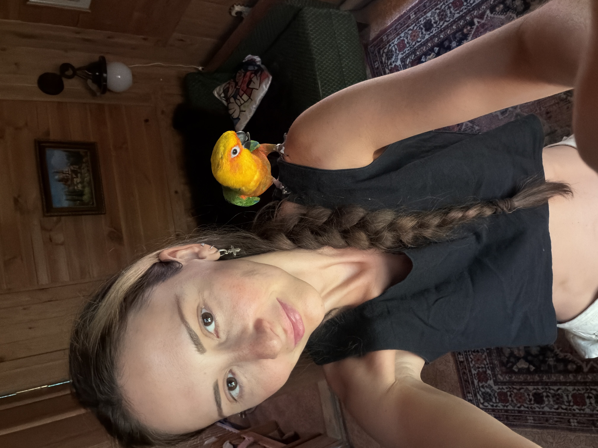 A woman with brown hair in a braid and a black tank top is taking a selfie. A small yellow and green parrot is perched on her left shoulder. The background shows a wooden-paneled room with a green couch and a rug on the floor. There is a framed picture on the wall and a light fixture. The image was taken in Udelnaya, Russia.