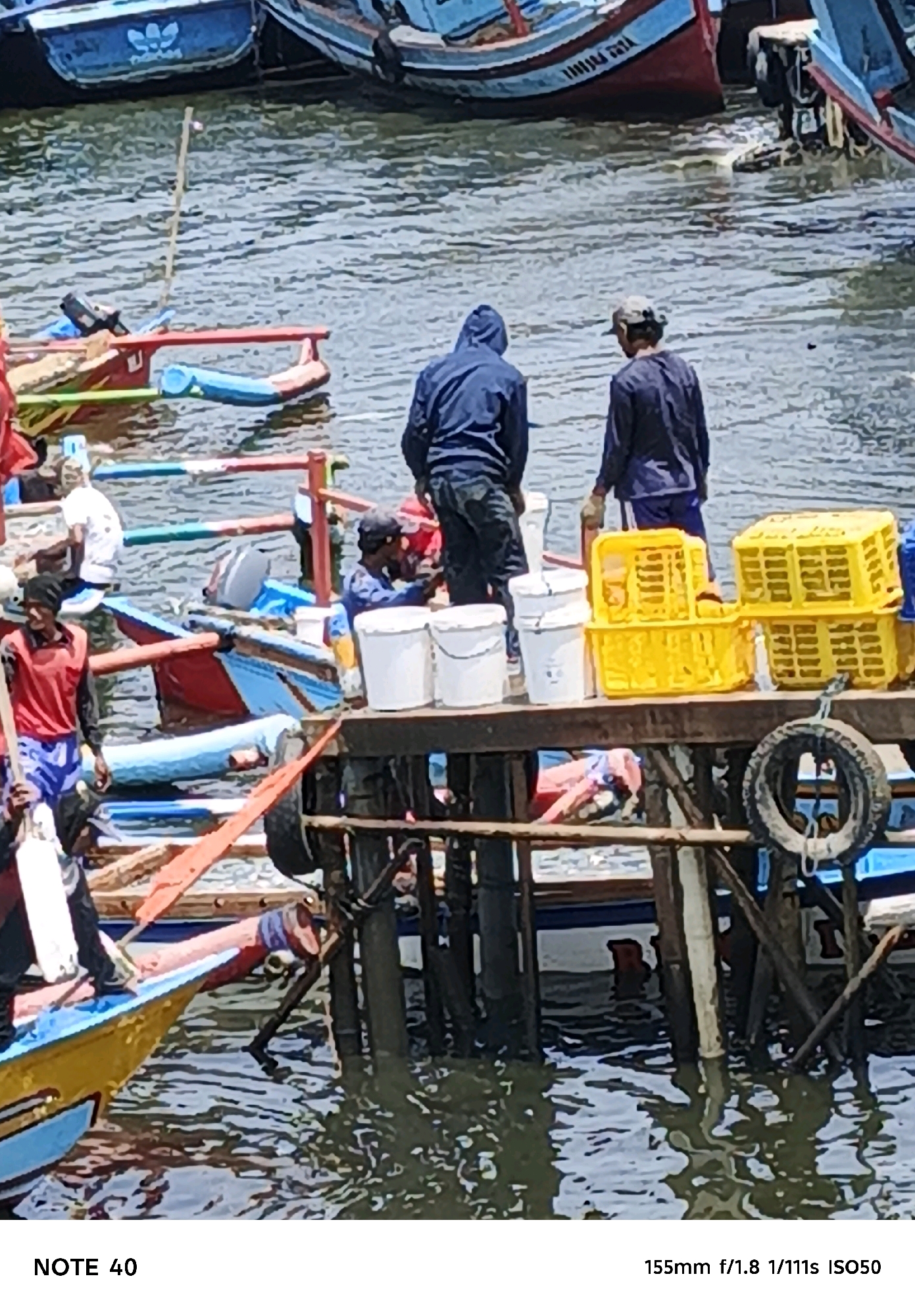 The image depicts a waterfront scene in Kelurahan Tegalkamulyan, Cilacap, Indonesia, likely a fishing harbor. In the foreground, a yellow and blue fishing boat is partially visible. Behind it, a wooden pier supports several individuals. Two men, one in a blue hooded jacket and another in a dark long-sleeved shirt and blue shorts, stand near the edge of the pier, looking towards the water. Another man in a red shirt and blue shorts stands on a boat to the left, holding a long object. Further back, yellow crates are stacked on the pier. The water is rippling, and other fishing boats, some blue and red, are moored in the background. The overall scene suggests activity related to fishing or boat maintenance.