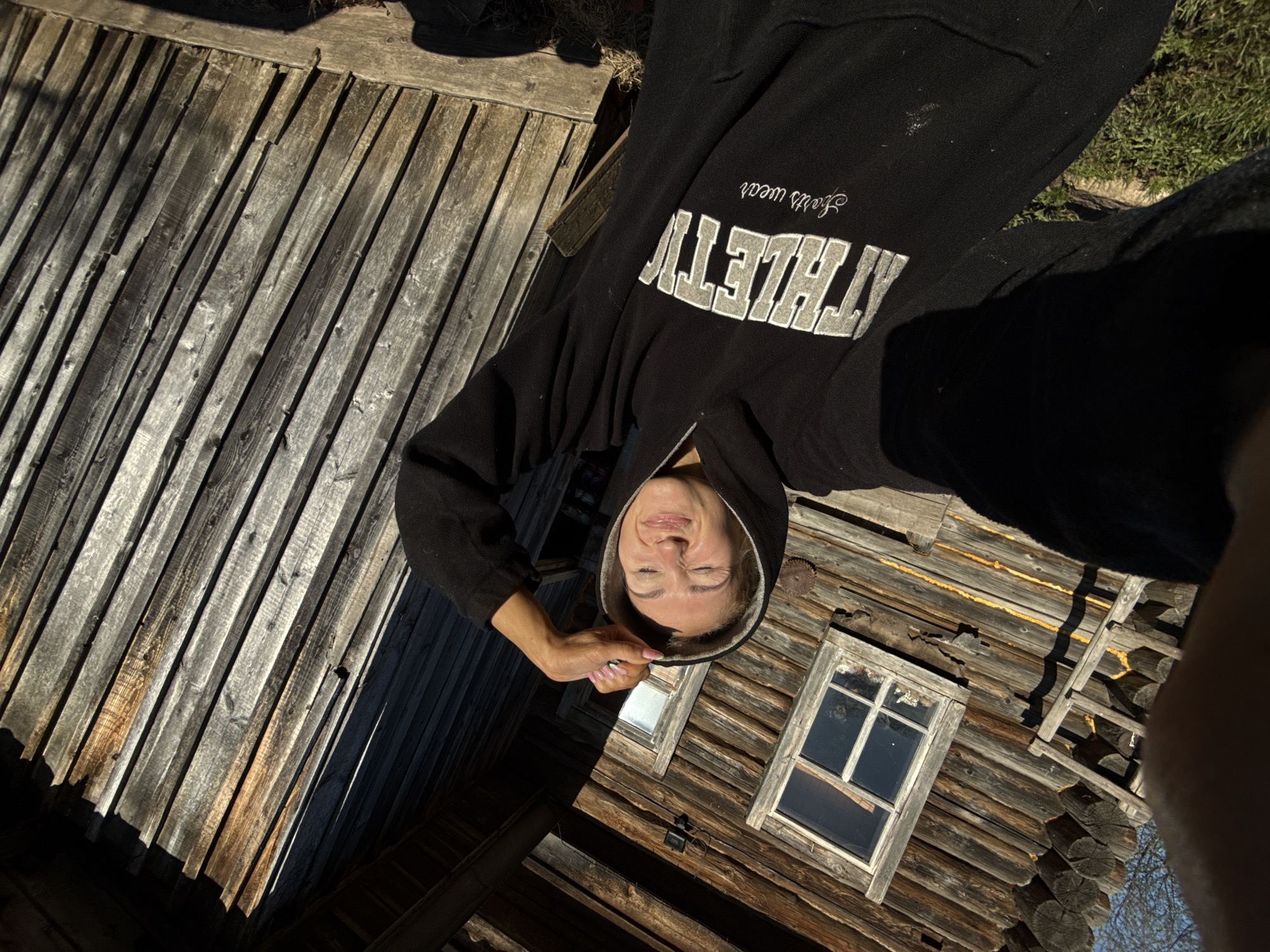 A low-angle shot shows a person in a black hoodie with "ATHLETIC" printed on the sleeve, standing in front of a wooden building with two windows. The background features a wooden fence and grass. The location is Nizhnyaya Chermoda, Russia.
