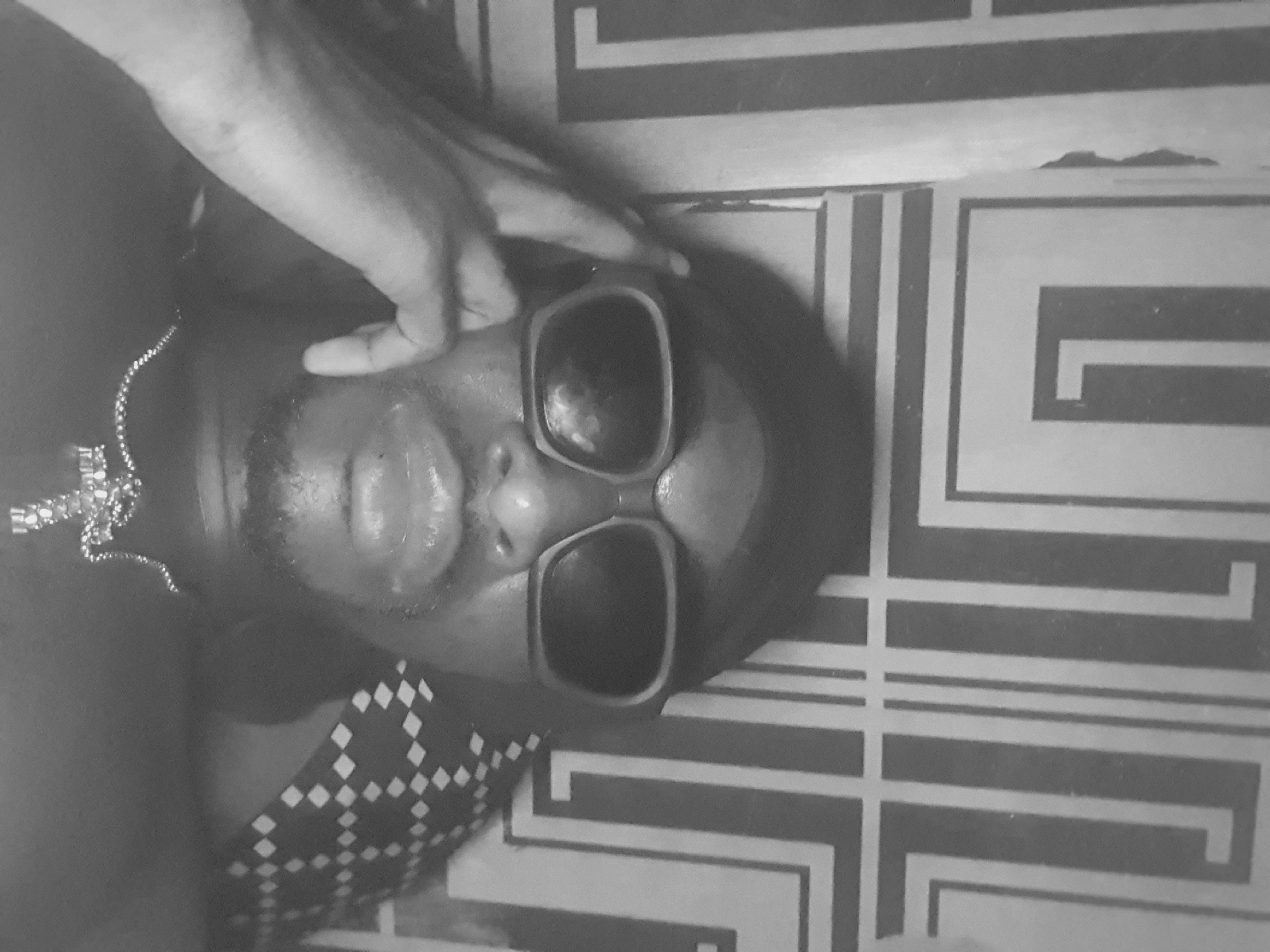 A black and white image of a person lying down wearing sunglasses. A hand touches their face. A necklace is visible. The person is wearing a patterned top. They appear to be on a surface with a linear geometric pattern. There is no information in the image to identify the location as Lokoja, Nigeria.
