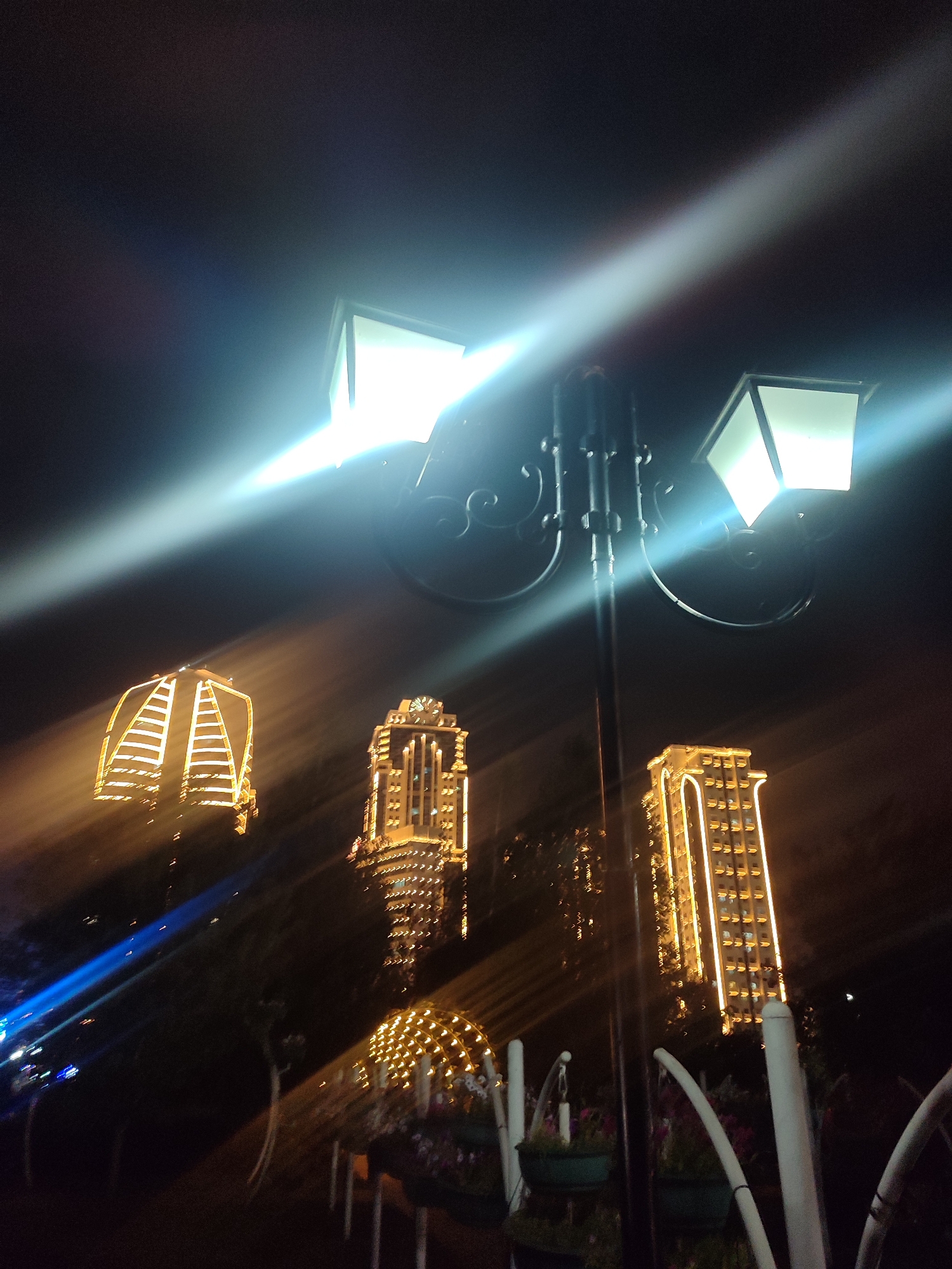 The image depicts a nighttime scene in Grozny, Russia, focusing on a streetlamp and illuminated buildings. The streetlamp in the foreground has two lamps, one of which is bright and emits strong light rays. In the background, three distinct buildings are visible, all adorned with numerous lights, creating a festive or illuminated architectural display. The leftmost building has a unique, stepped pyramid-like shape. To its right is a taller, more rectangular building with a clock face near its peak. The building furthest to the right is also tall and rectangular. The lighting in the scene is dynamic, with light beams from the streetlamp stretching across the frame, and the buildings glowing brightly against the dark night sky. The image appears to have been taken with a camera that was in motion during the exposure, resulting in motion blur and light streaks. The environment suggests an urban setting, likely a public space or a prominent area within the city. There are no people visible in the image.