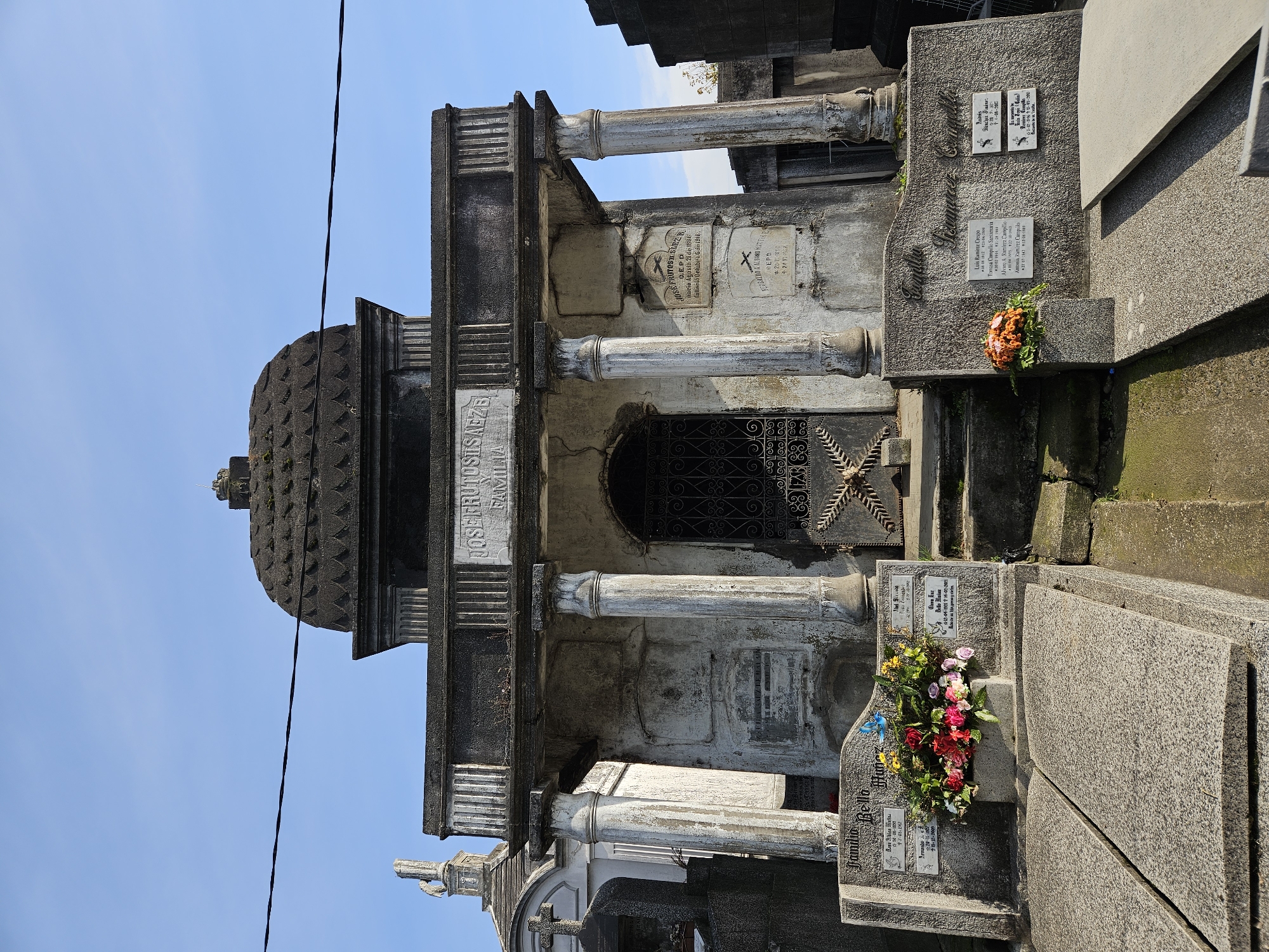 This image shows a mausoleum within a cemetery. The structure is made of weathered stone and features a dome-shaped roof, columns, and an arched entrance with an iron gate. Plaques with inscriptions are visible on the mausoleum's facade and on surrounding grave markers, adorned with flowers. The scene is likely taken in a cemetery in Osorno, Chile, based on the inscriptions written in Spanish on the gravestones. A utility wire cuts diagonally across the upper left of the frame.