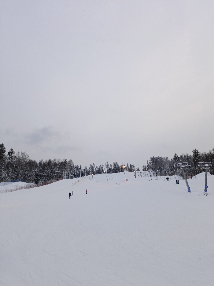 The image captures a wide view of a snow-covered ski slope under an overcast sky. The upper two-thirds of the frame are filled with a uniform, light grey sky, indicating a heavily clouded or overcast winter day, possibly nearing dusk given the artificial lights visible.
The lower third of the image is dominated by the expansive white snow of the ski slope, which appears to be groomed, though faint ski tracks are visible across its surface. Along the horizon, a dense line of trees, heavily dusted with snow, separates the sky from the ski area. This treeline consists of both dark evergreen conifers and some bare deciduous trees.
Various infrastructure typical of a ski resort is visible. On the right side of the slope, several prominent ski lift towers with blue accents stand against the snow. Further up the slope, extending towards the center, are multiple smaller pull-type ski lifts, likely T-bars or button lifts, with their support structures and cables visible. Artificial lights on poles are illuminated across different sections of the slope, casting a subtle glow, particularly around the lift areas. An orange safety net or fencing runs along the left edge of the groomed slope.
Several people are scattered across the slope, engaged in winter activities. Towards the left-middle of the slope, a person in dark clothing is standing on skis or a snowboard, accompanied by another individual in lighter attire nearby. Further down and to their right, a distinct figure in a bright red ski suit is visible, appearing to be skiing or standing. On the right side of the slope, closer to the large ski lift towers, a few small groups of people are seen standing or walking on the snow, possibly waiting for lifts or congregating.
The overall atmosphere is quiet and serene, characteristic of a winter day at a ski resort, with the soft, diffused light from the sky and the illuminated artificial lights providing visibility. There is no visible text in the image.